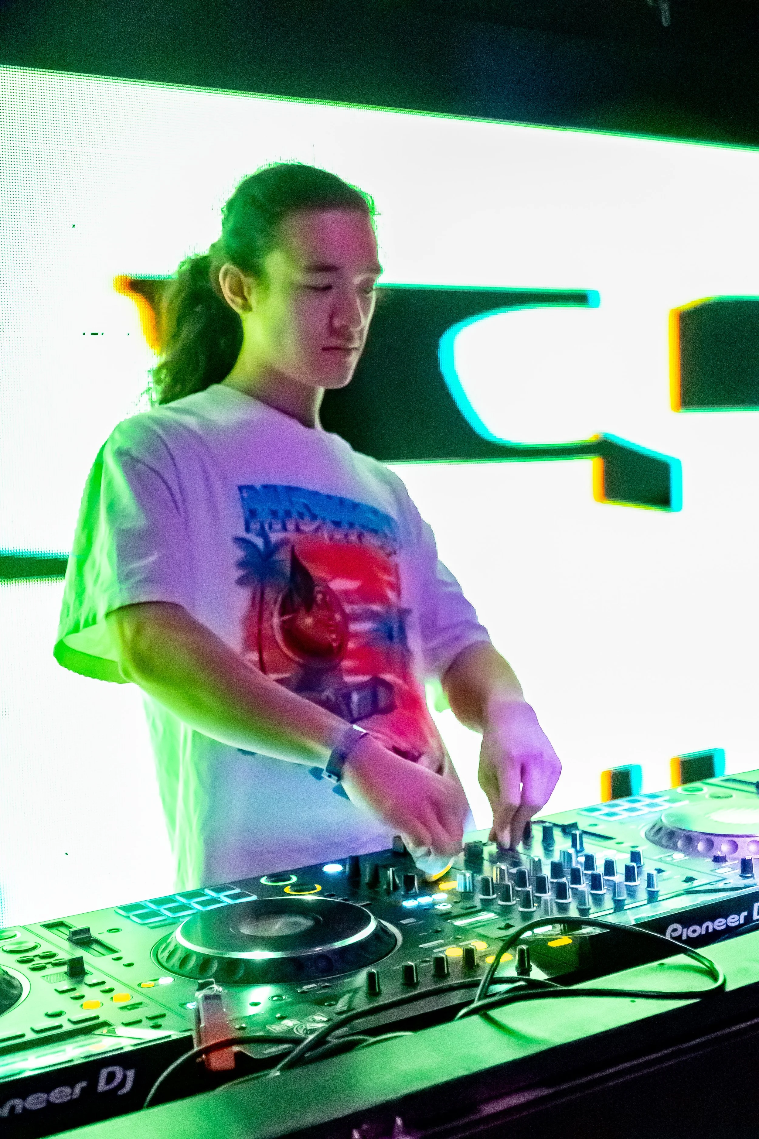 A young person with long hair tied back, wearing a graphic T-shirt, DJing with Pioneer DJ equipment in front of a vibrant, colorful LED screen.