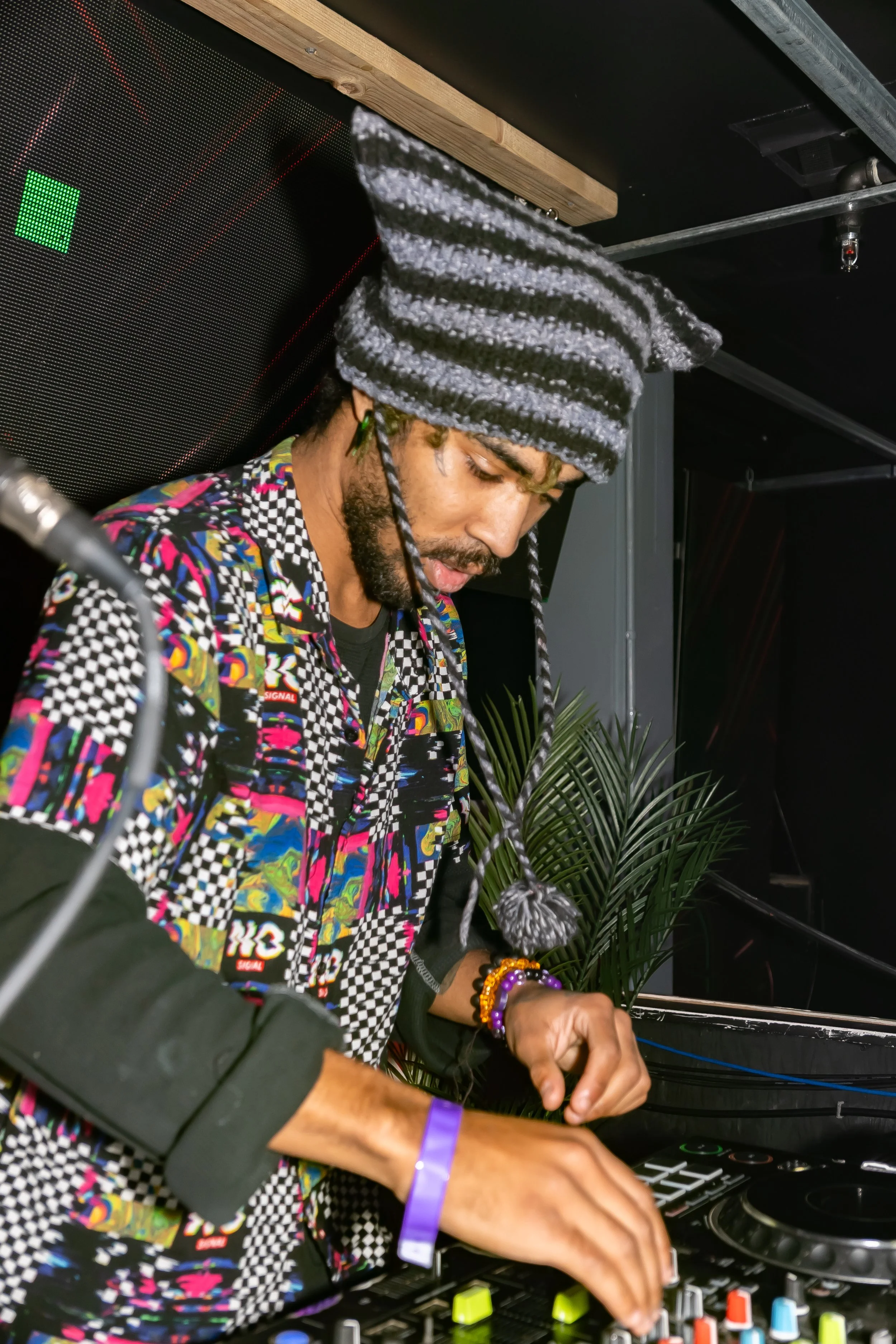 A DJ wearing a colorful patterned shirt and a striped gray beanie hat, focused on mixing music on a DJ controller with his hands, in a nightclub setting with greenery in the background.