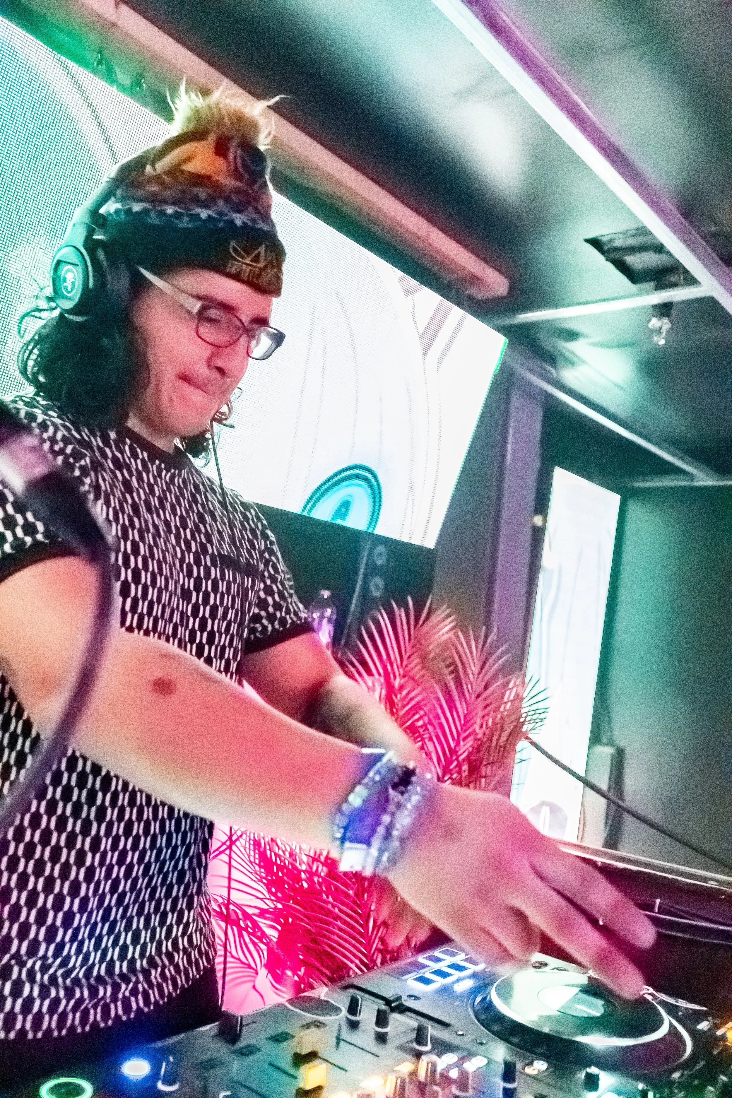 A woman DJ with curly dark hair, glasses, wearing headphones and a patterned black-and-white shirt, is mixing music at a nightclub, with colorful lights and screens in the background.