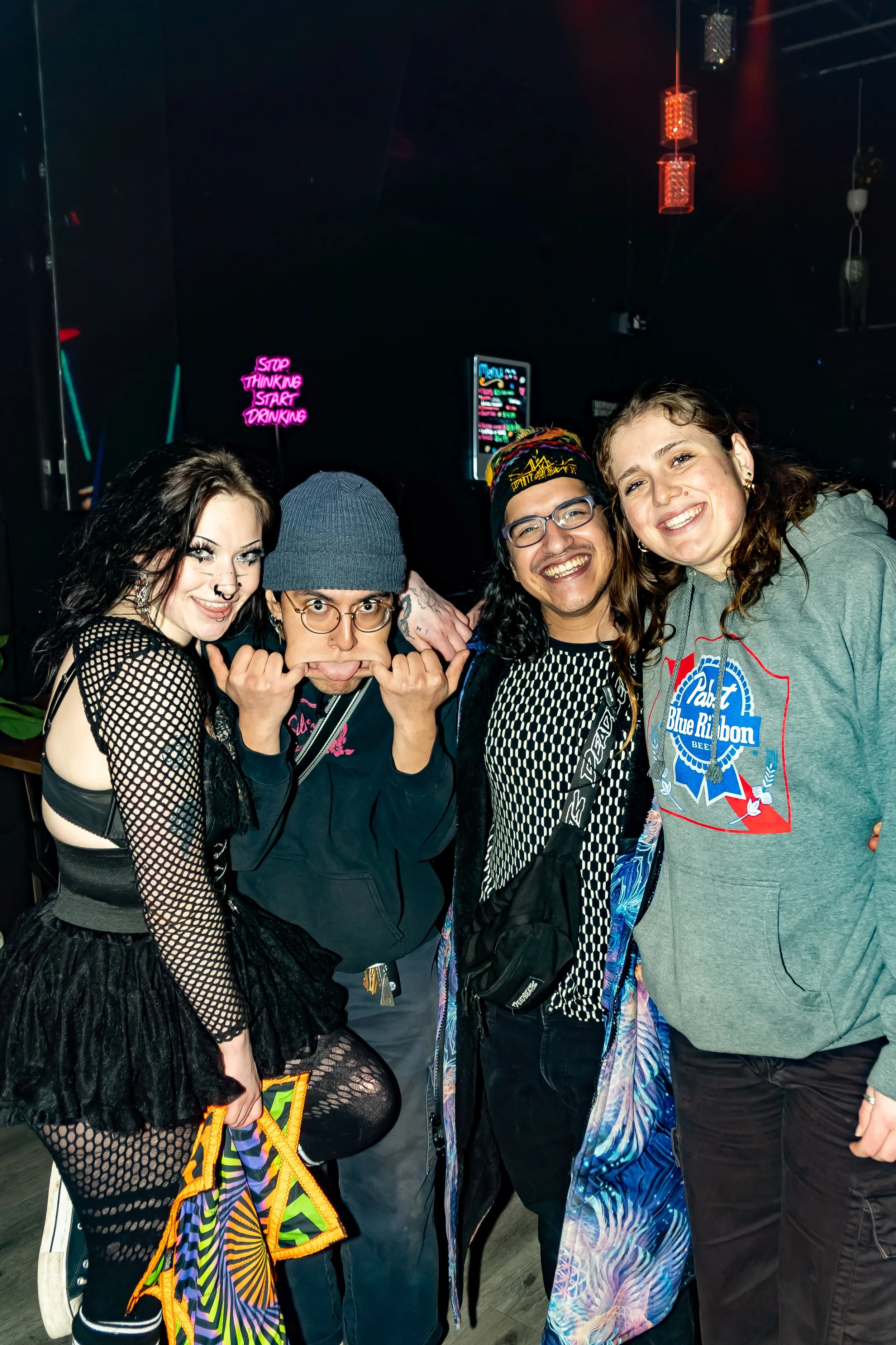 Four friends posing together at a nightclub or bar, with a dark background and neon signs visible