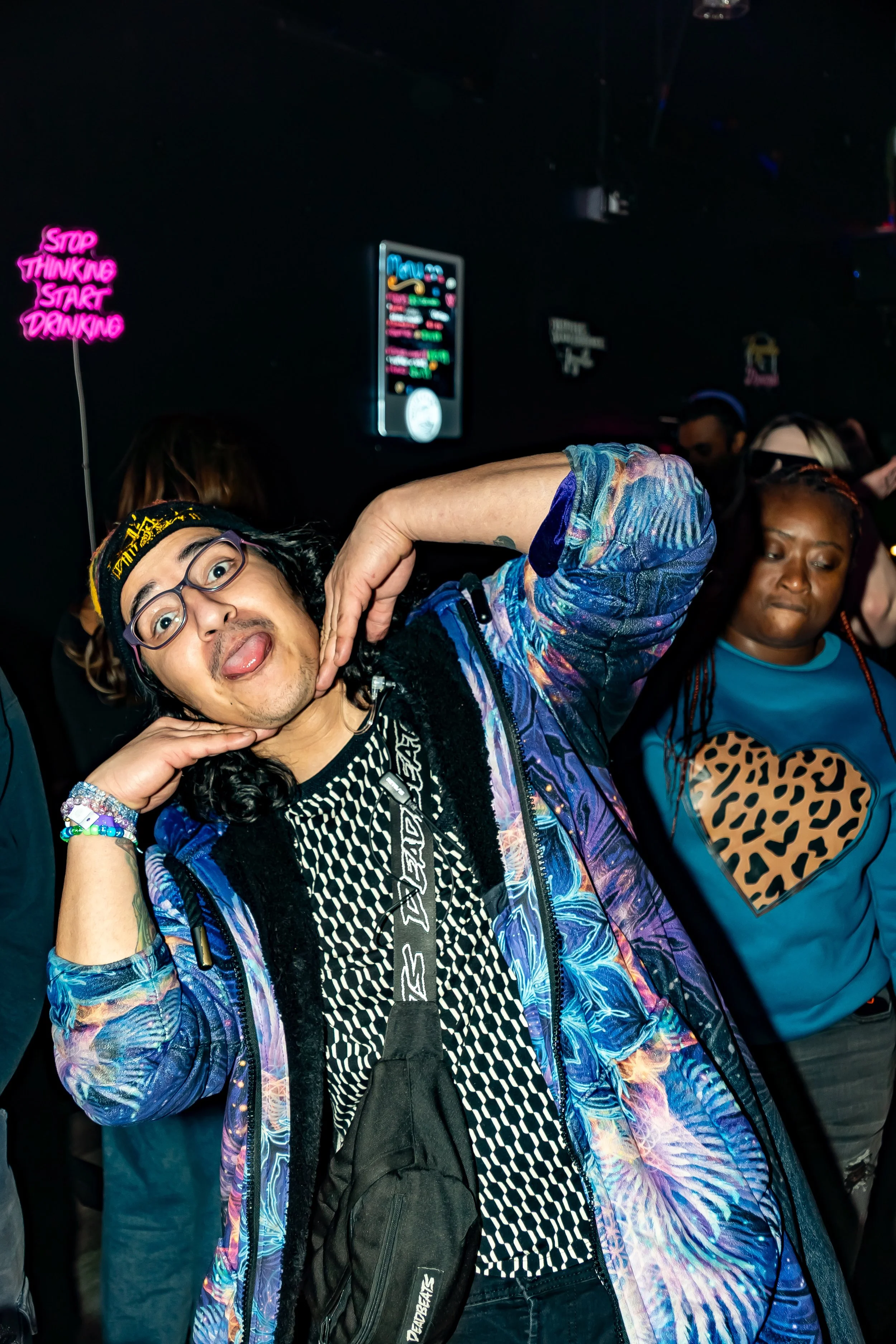 Person with glasses and long hair playfully poses with tongue out, hands framing face, in a crowded club with neon signs and other people dancing.