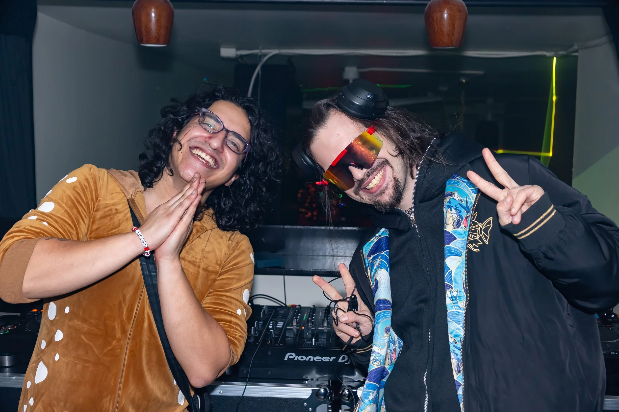 Two smiling DJs in a nightclub, one wearing glasses and a brown jacket, the other wearing headphones, sunglasses, and a black jacket, with DJ equipment in the background.