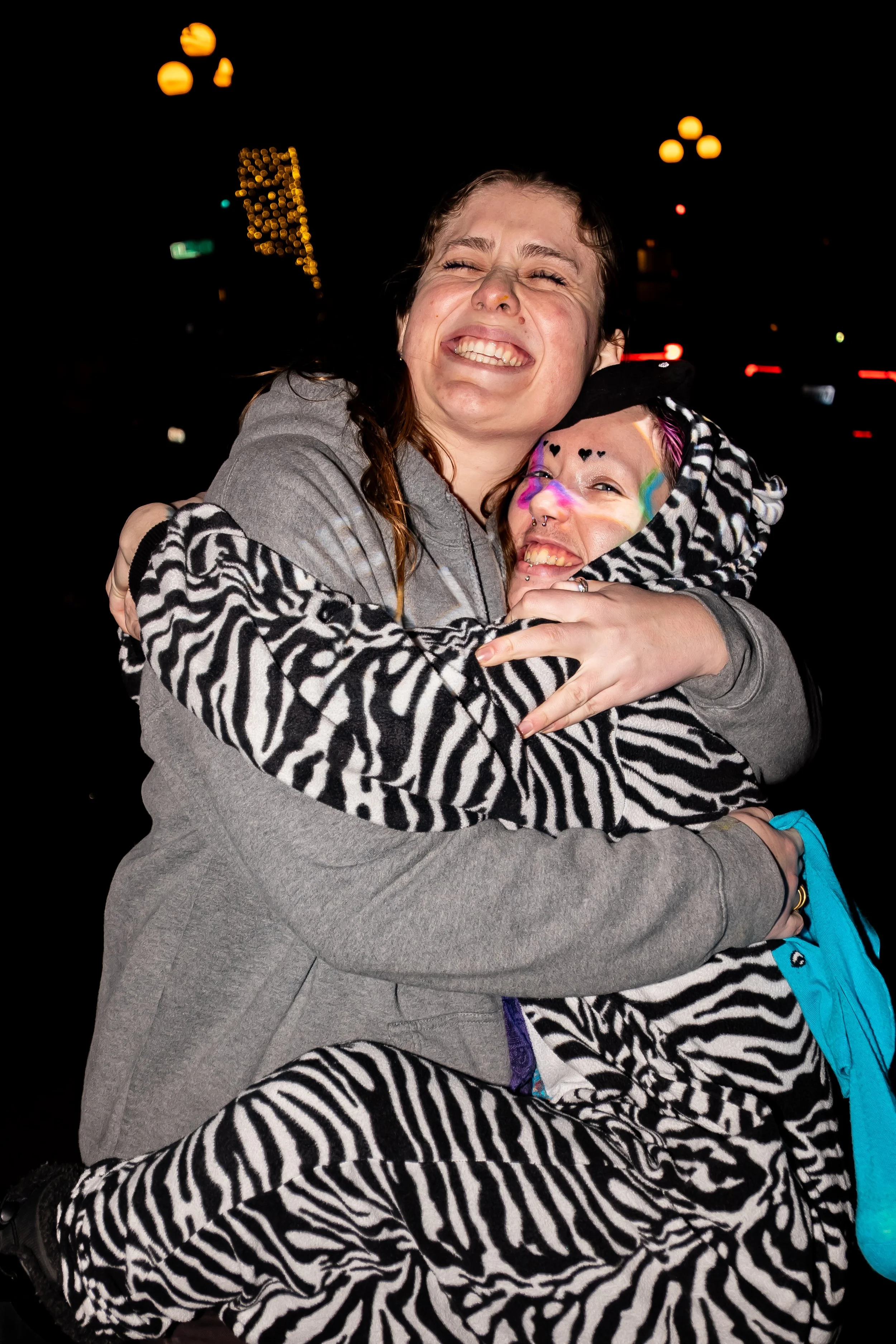 Two young women hugging at night, one with face painted and colorful accessories, both smiling, in zebra-patterned outfits.