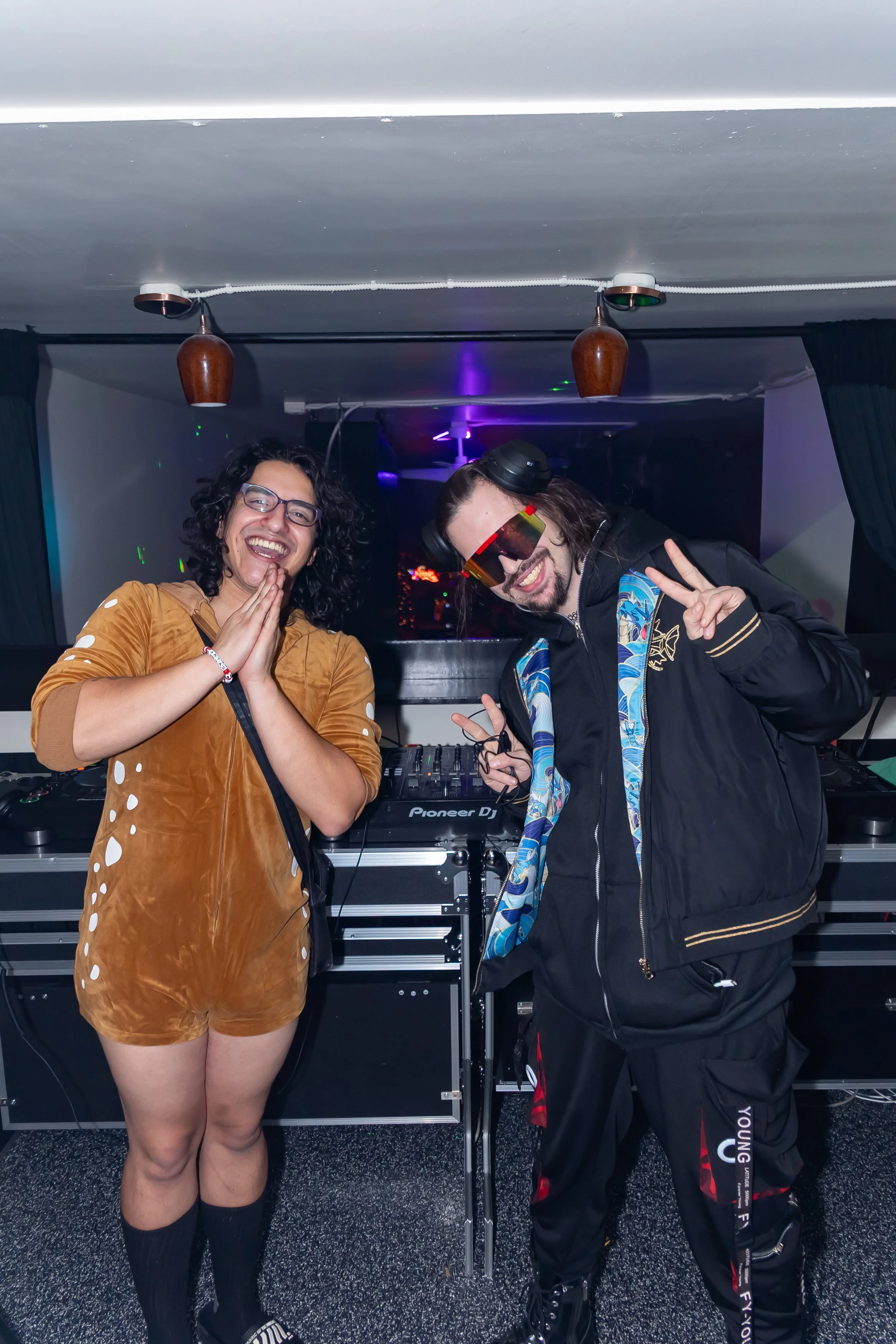 Two people smiling in a nightclub, standing in front of DJ equipment. One person is wearing a brown outfit and glasses, the other is wearing a black jacket with colorful patterns, sunglasses, and headphones.