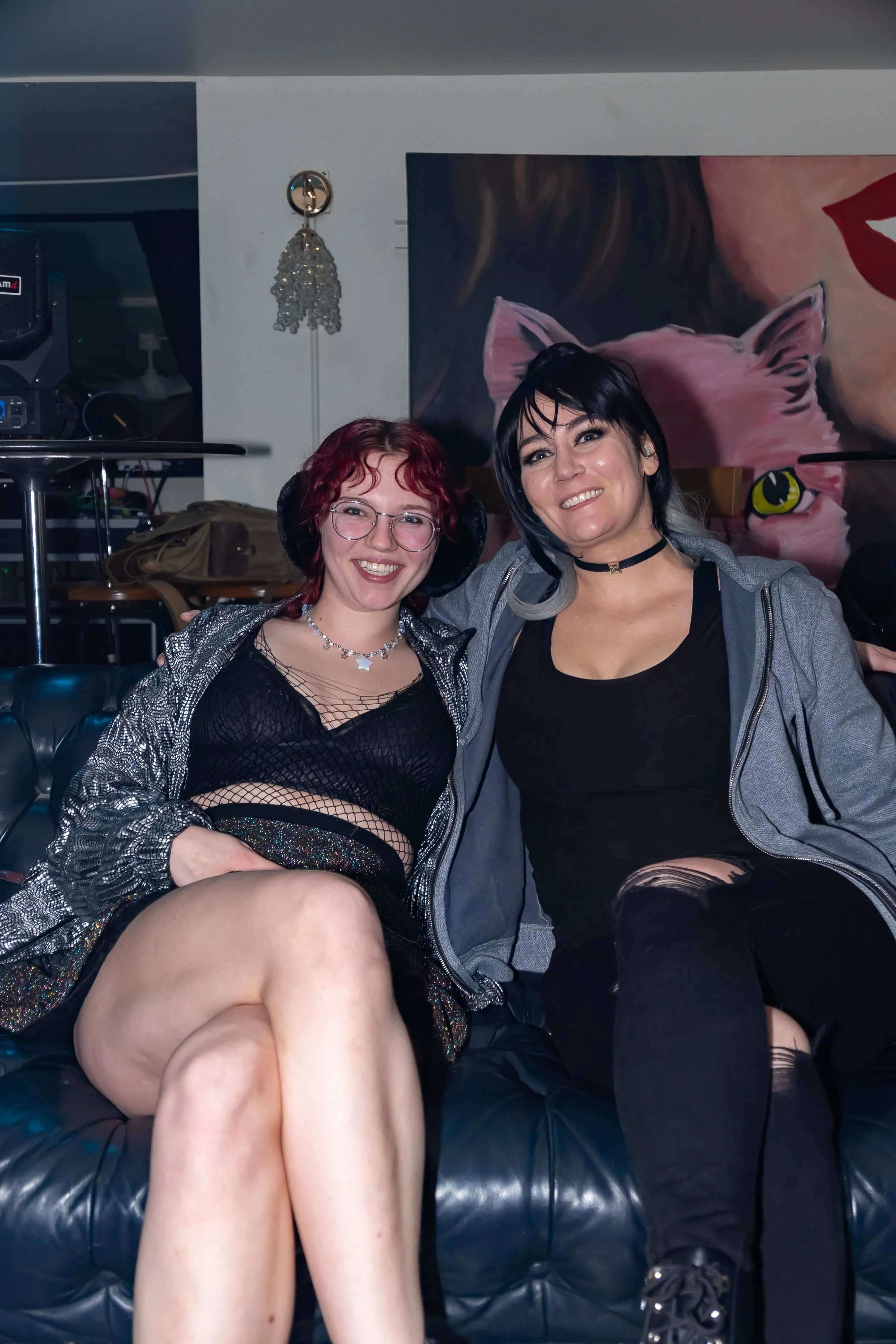 Two women sitting on a black leather couch, smiling at the camera. One has red curly hair, glasses, and headphones, wearing a black fishnet top and sparkly shorts. The other has black hair with gray tips, wearing a black top, gray jacket, and ripped 