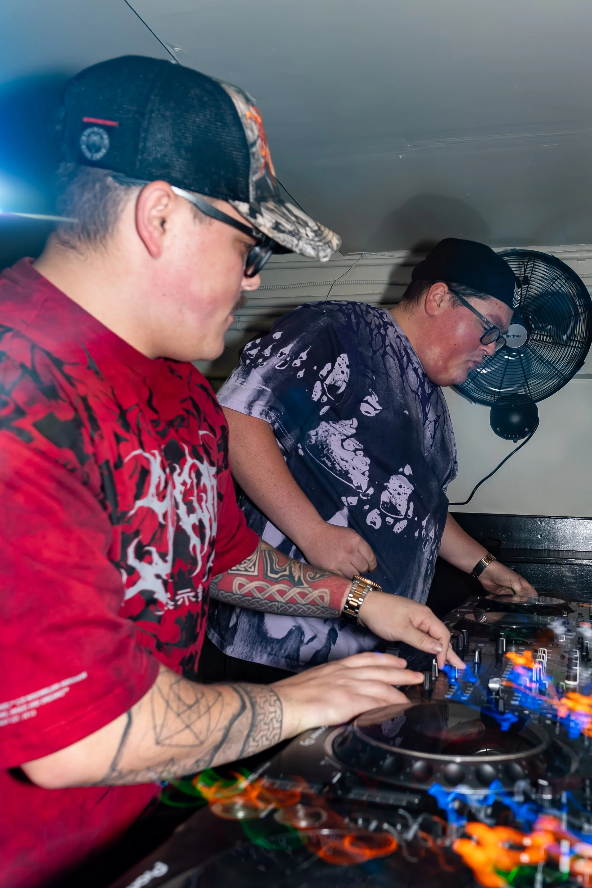 Two young men DJing with mixer and turntables in a dimly lit room, one wearing a red T-shirt and the other in a tie-dye T-shirt, both focused on their equipment.