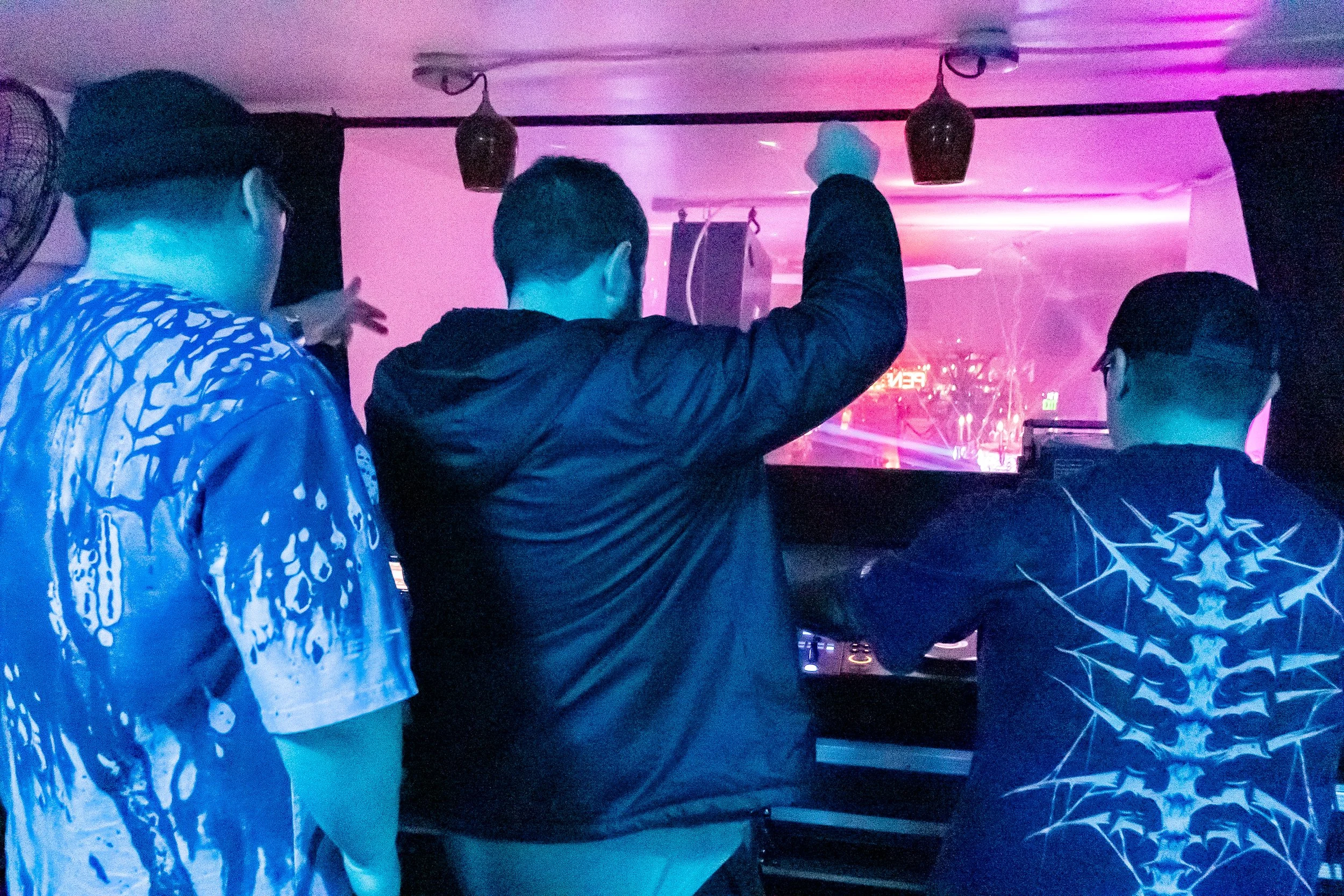 Three young men are standing in front of a DJ booth, with one adjusting equipment. The scene features colorful neon and purple lighting, creating a lively atmosphere.