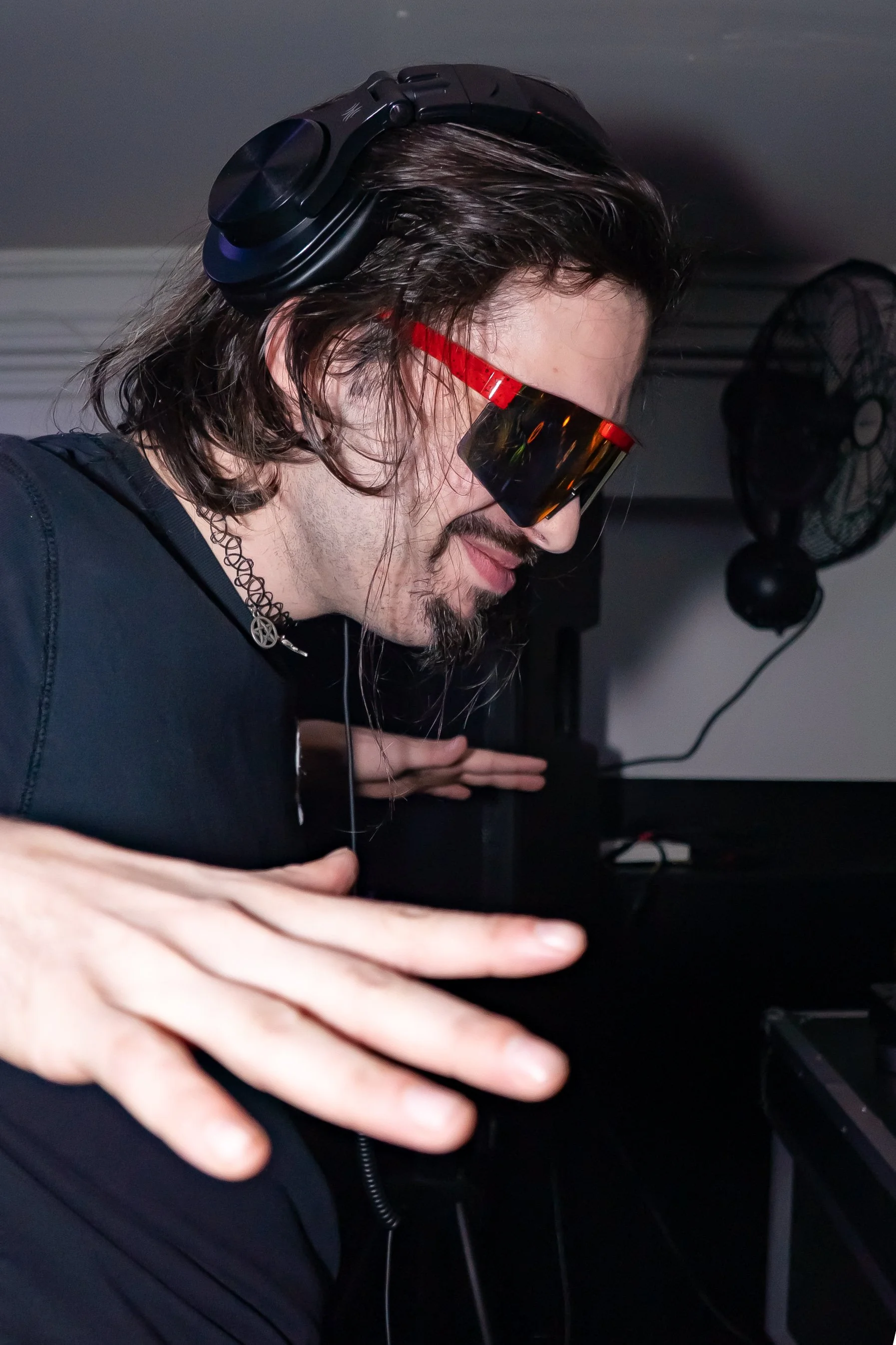 A man with long hair and a goatee wearing sunglasses, headphones, and a choker, appears to be DJing or mixing music in a dimly lit room.