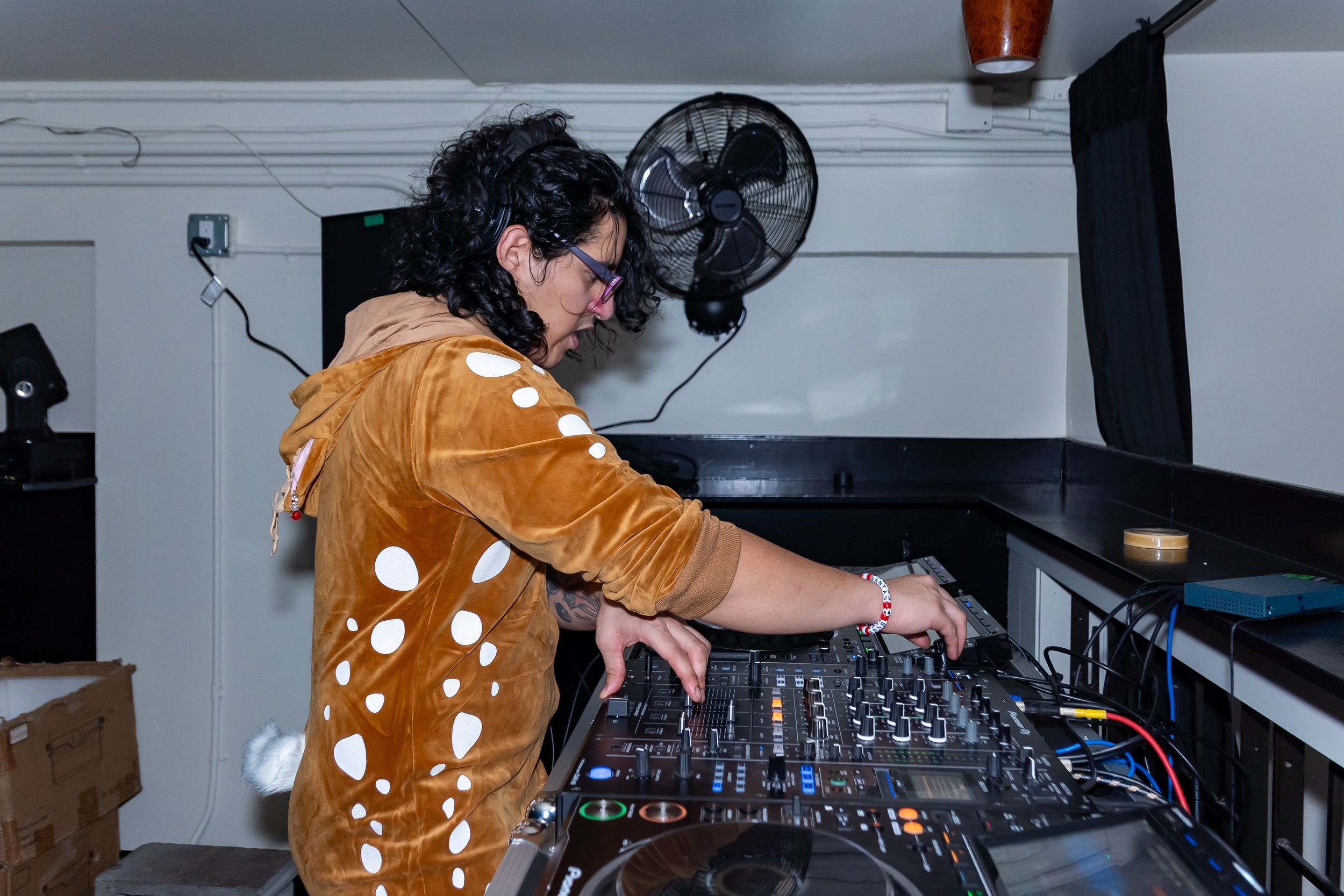 A woman with curly black hair, wearing glasses, a brown hoodie, and a red and white bracelet, is DJing with a professional DJ mixer and turntables in a dark room with a black curtain on the right, a fan on the wall, and a small box on the counter.