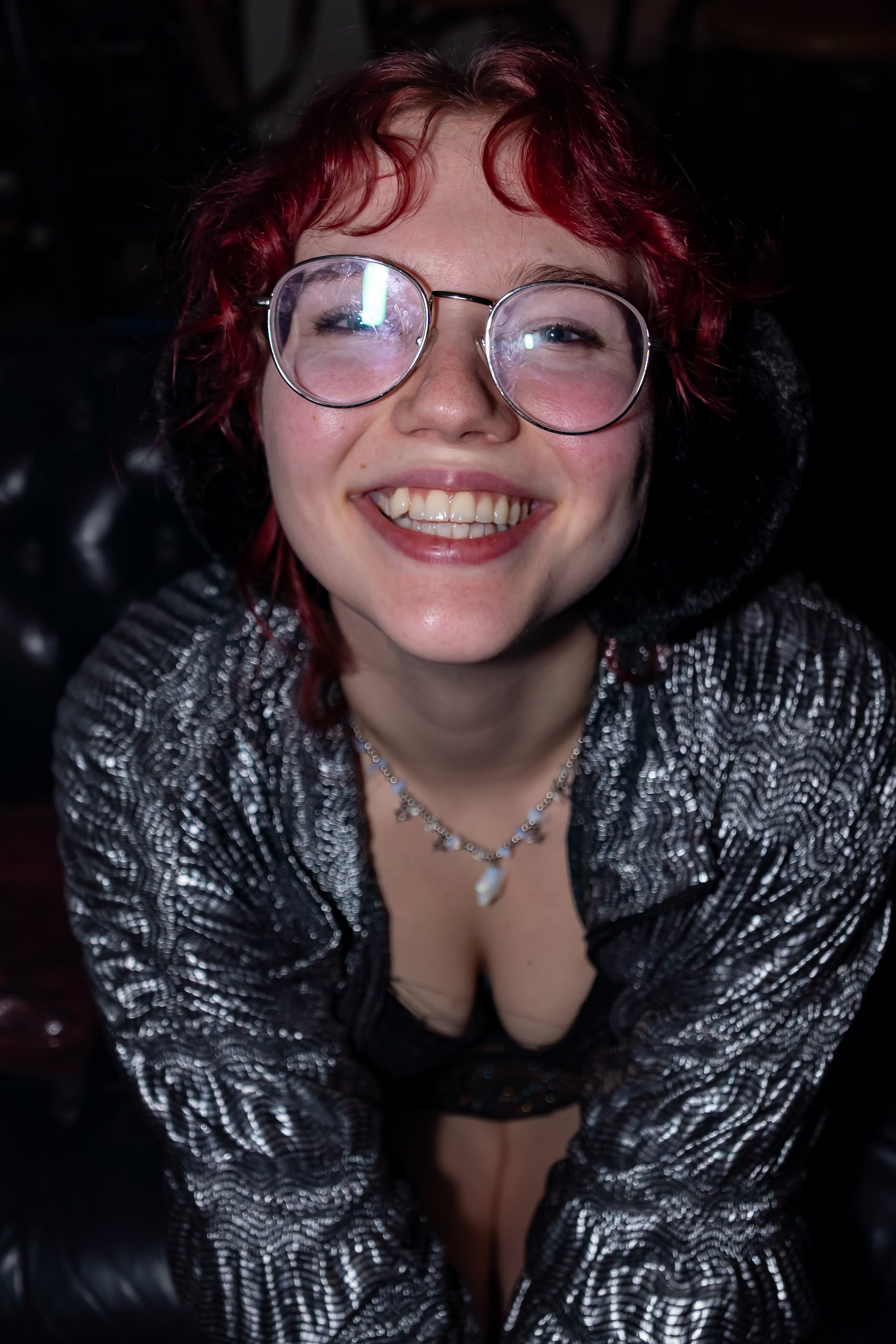 A young woman with red curly hair and glasses smiling at the camera, wearing a patterned shiny top and a necklace.