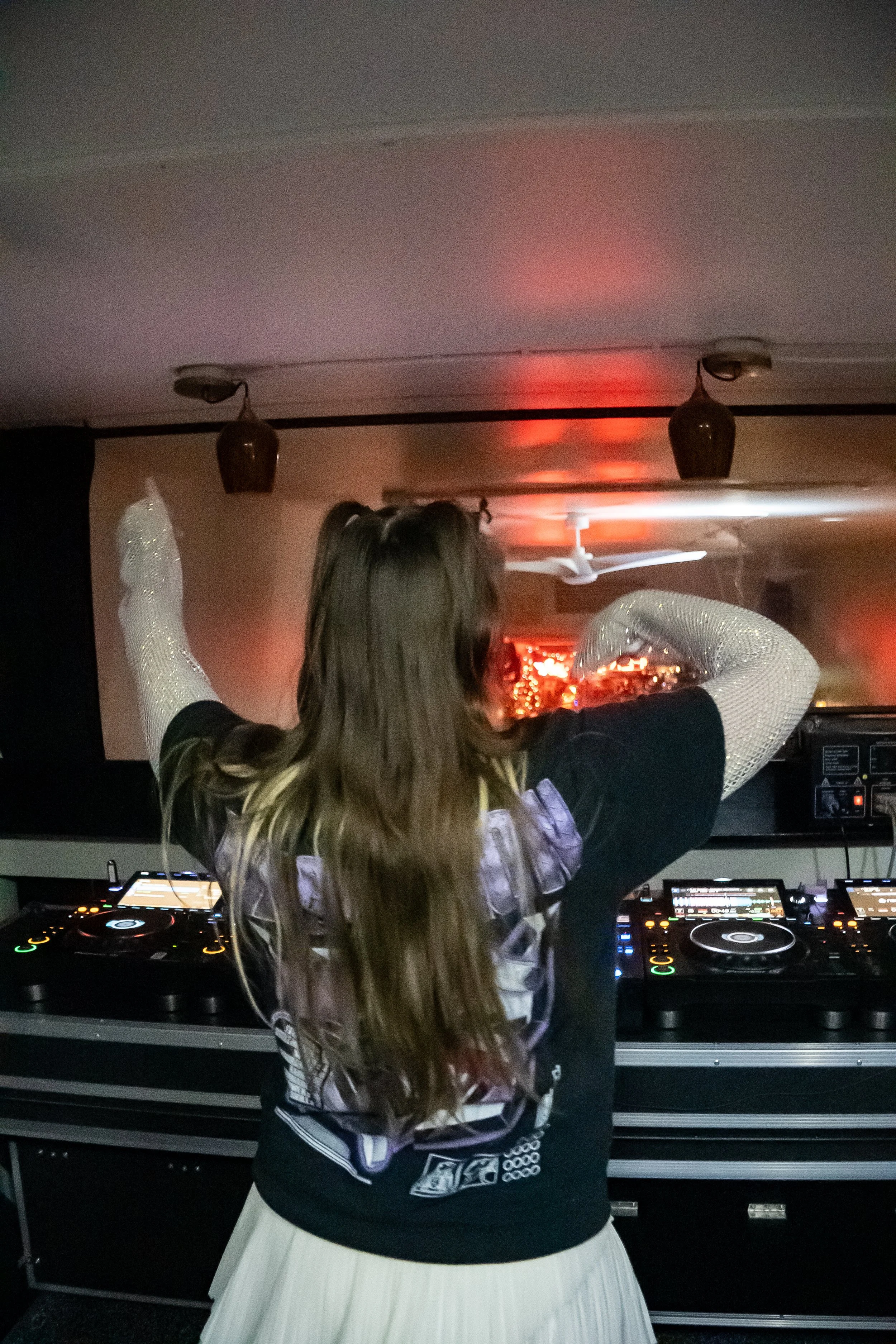 A woman with long, wavy hair and glittery arm sleeves DJing at a club or party, facing a DJ setup with turntables and mixing equipment, with red and orange lighting in the background.