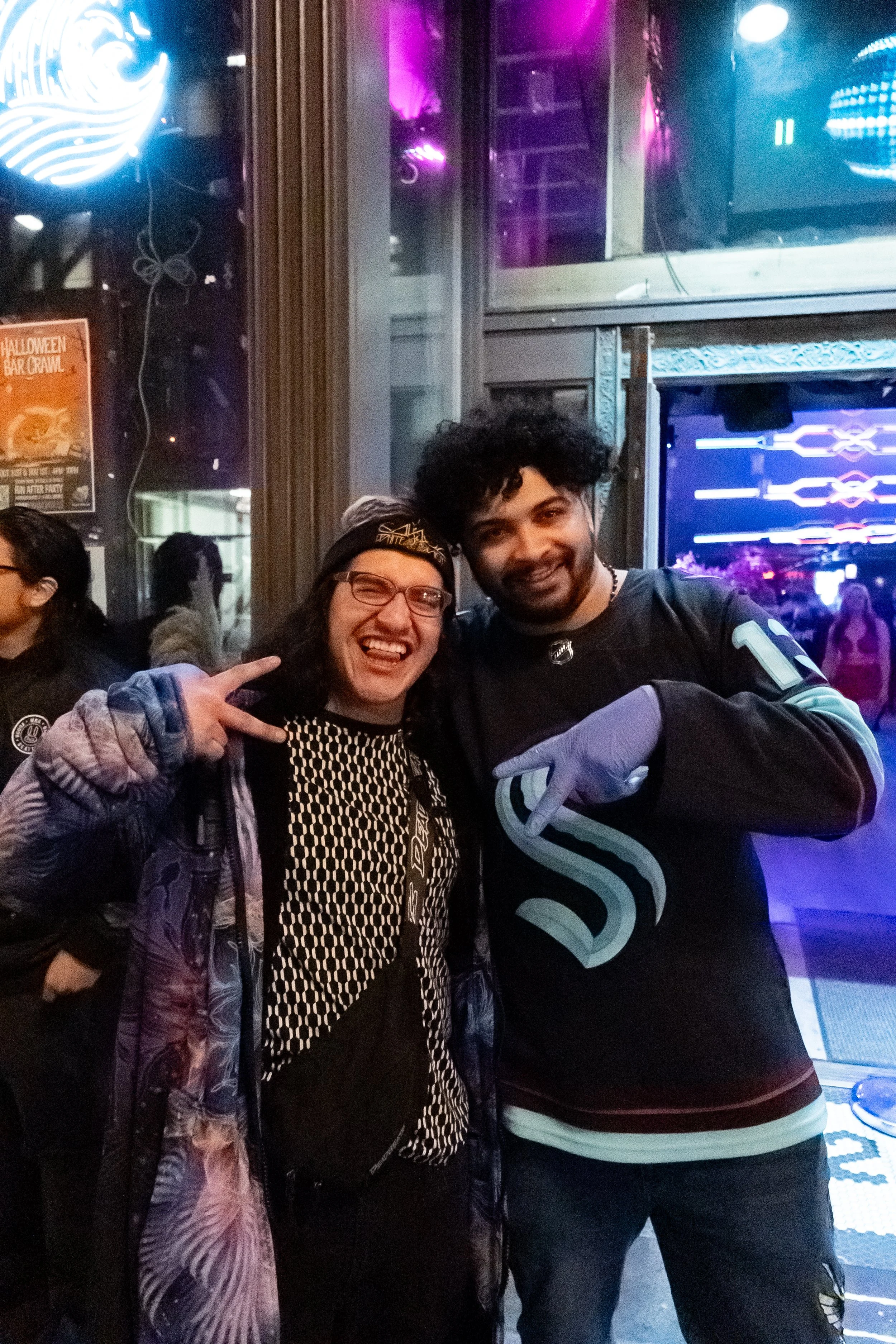 Two people smiling and posing for a photo inside a nightclub, one woman making a peace sign, the man pointing at her, neon lights and other individuals in the background.