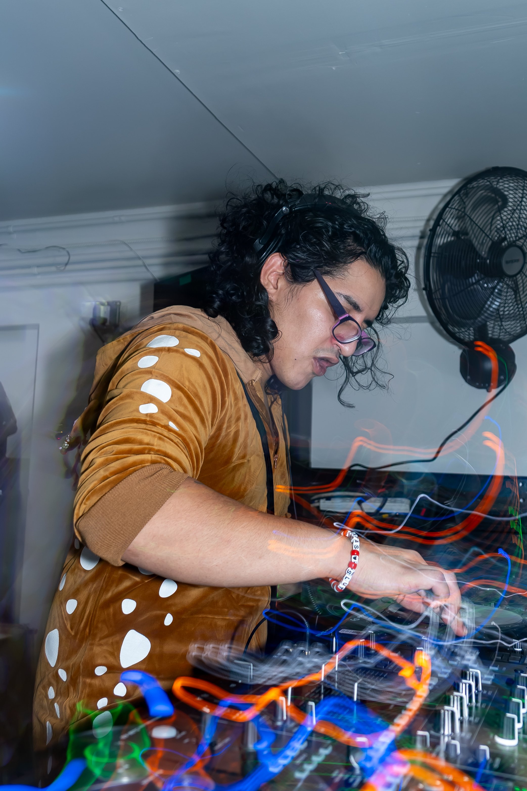 A DJ with curly black hair, glasses, and a brown hoodie with white polka dots, mixing music on electronic DJ equipment with colorful light trails, in a room with a fan and wall-mounted items.