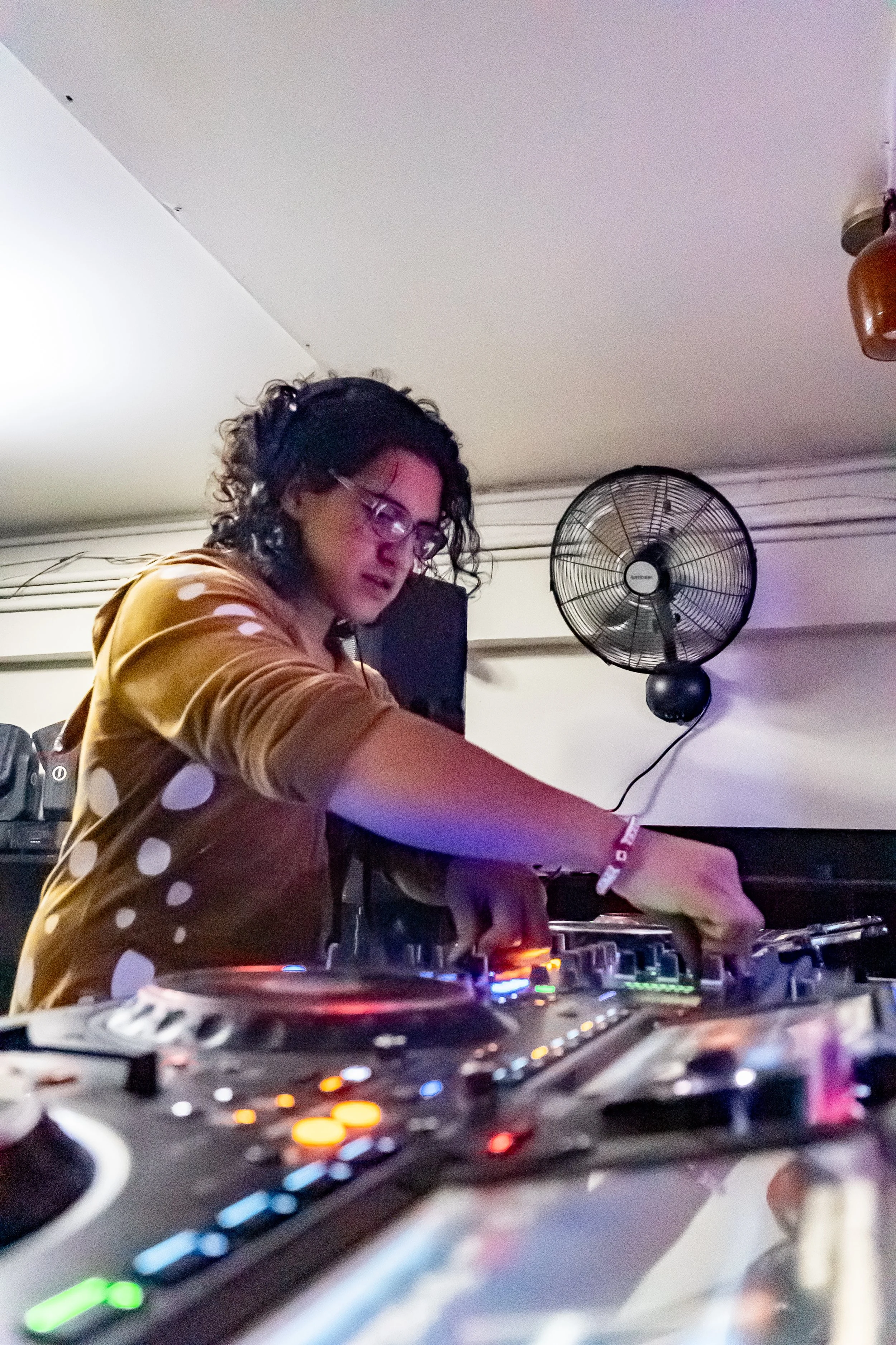 A woman with curly dark hair and glasses, wearing a brown shirt with white polka dots, is DJing with headphones in a room with a wall fan behind her.