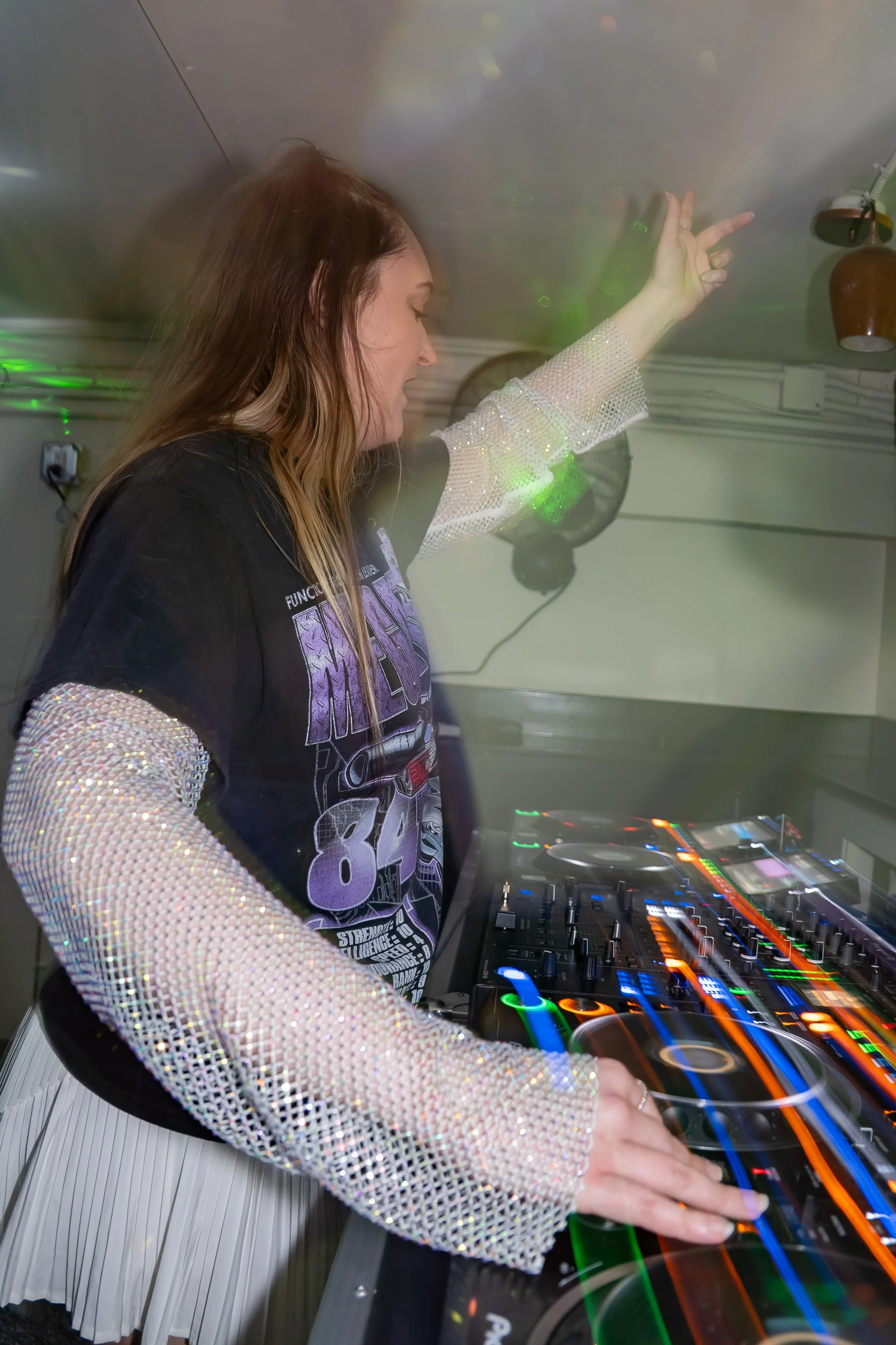 A female DJ with long brown hair, wearing a black T-shirt with colorful graphics and shiny, glittery, long-sleeve top, is playing music with DJ equipment in a club or party setting.