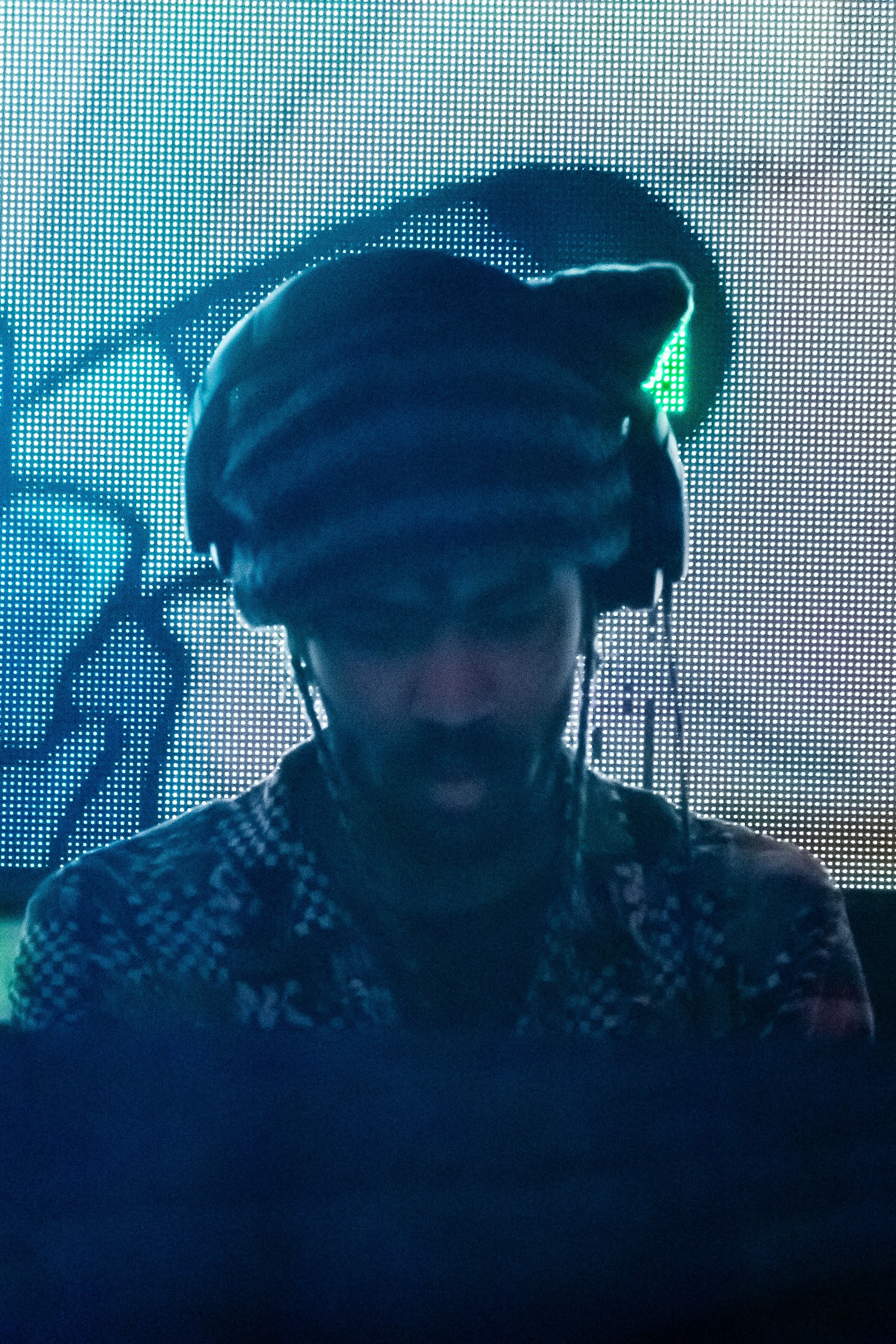 A person wearing headphones and glasses, standing in front of a green lit LED screen, with a mural or digital artwork in the background.