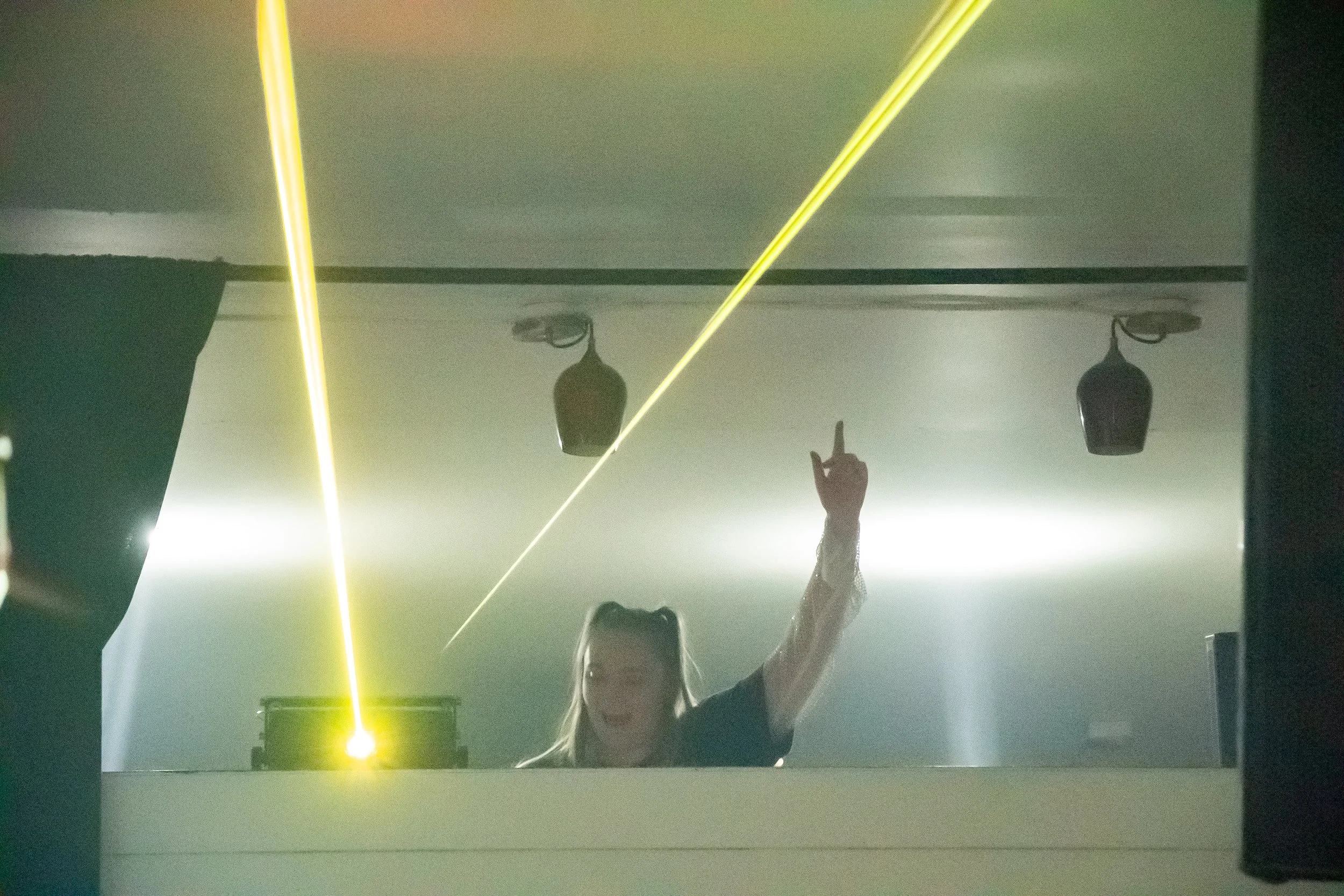 A female DJ or performer with blonde hair in pigtails, raising her right hand with her index finger pointing upward, in a dimly lit club with yellow laser lights and fog, behind a DJ booth.