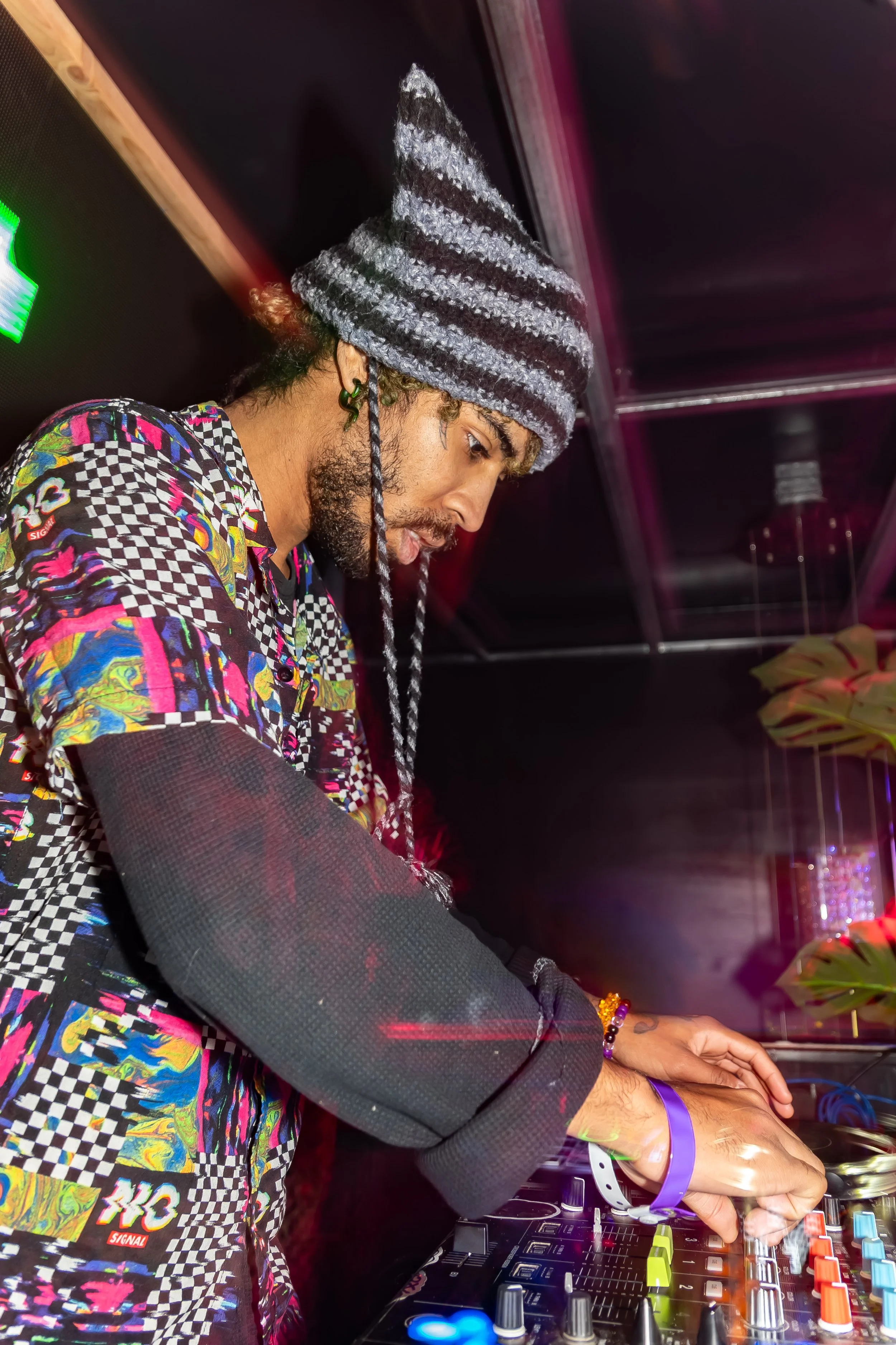 A DJ wearing a gray and black striped beanie and a colorful patterned shirt is mixing music on a DJ console in a dimly lit nightclub or party setting.
