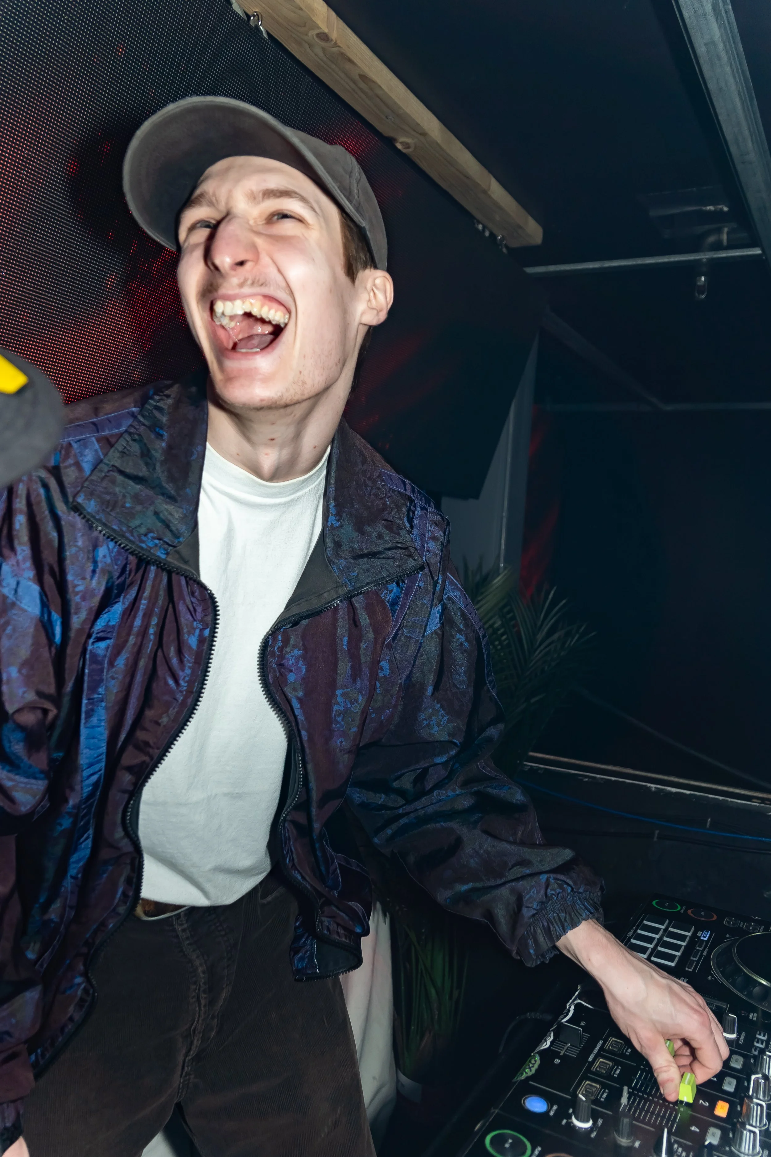 A young man wearing a gray cap, a white t-shirt, and a shiny, dark windbreaker is smiling widely while DJing at a party, with DJ equipment in front of him and a dark background.