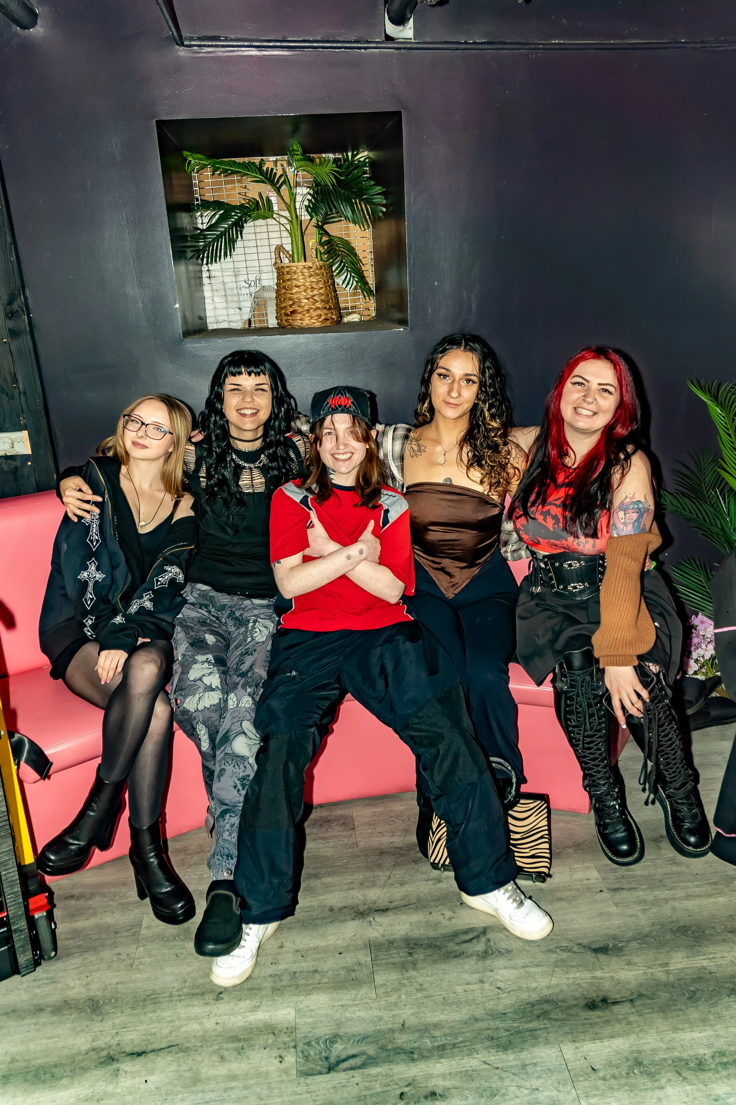 A group of five women sitting on a pink bench in a stylish indoor setting, with a dark wall and a potted plant in the background.