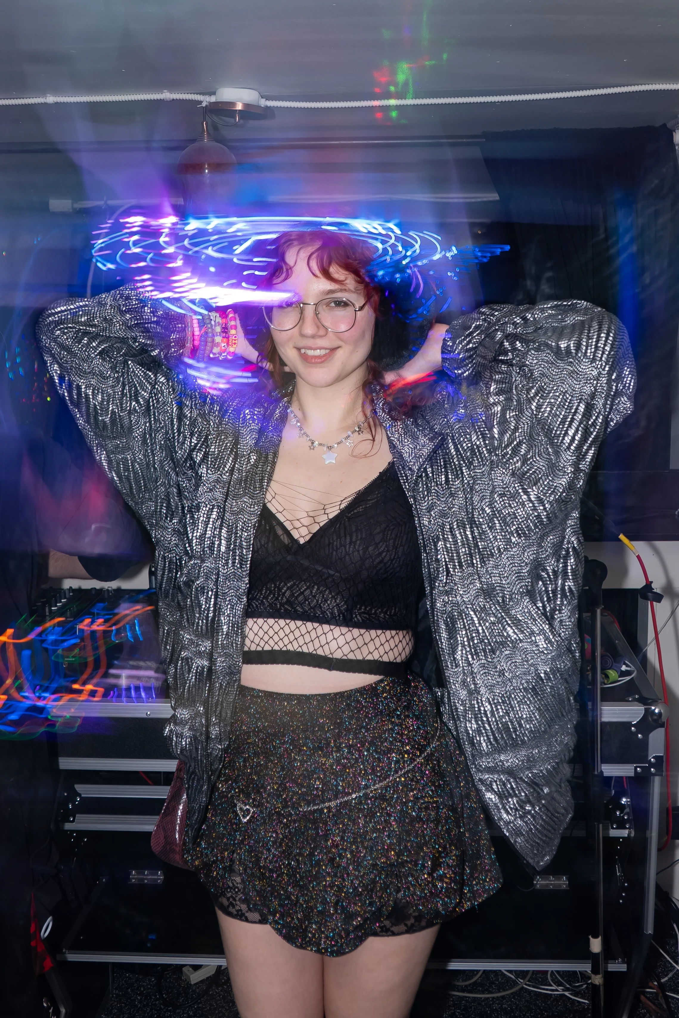 A young woman with red hair and glasses dancing at a party with colorful lights and a DJ setup in the background.