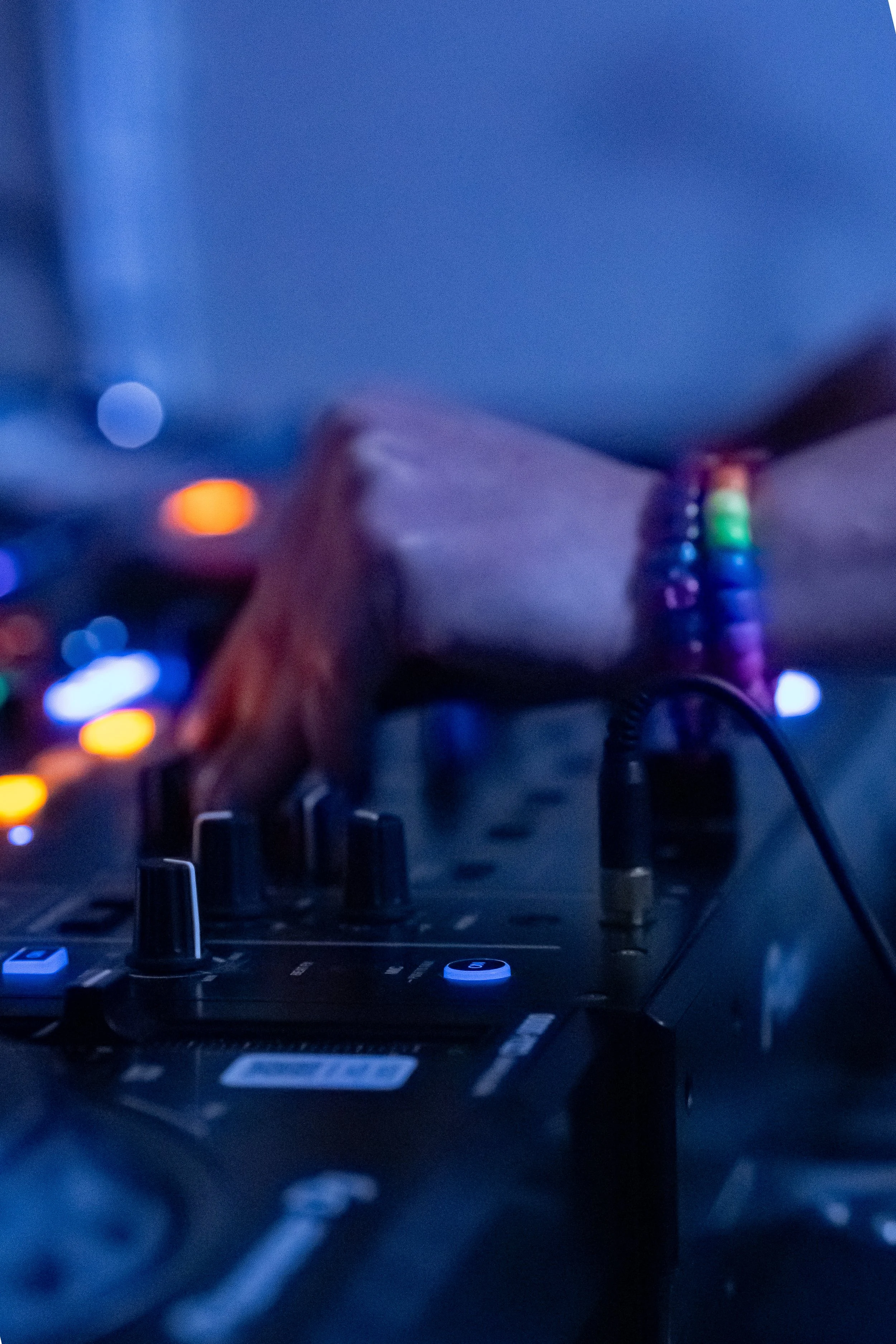 Close-up of DJ equipment with knobs and faders, a hand wearing rainbow-colored beaded bracelet adjusting controls, and colorful blurred lights in the background.