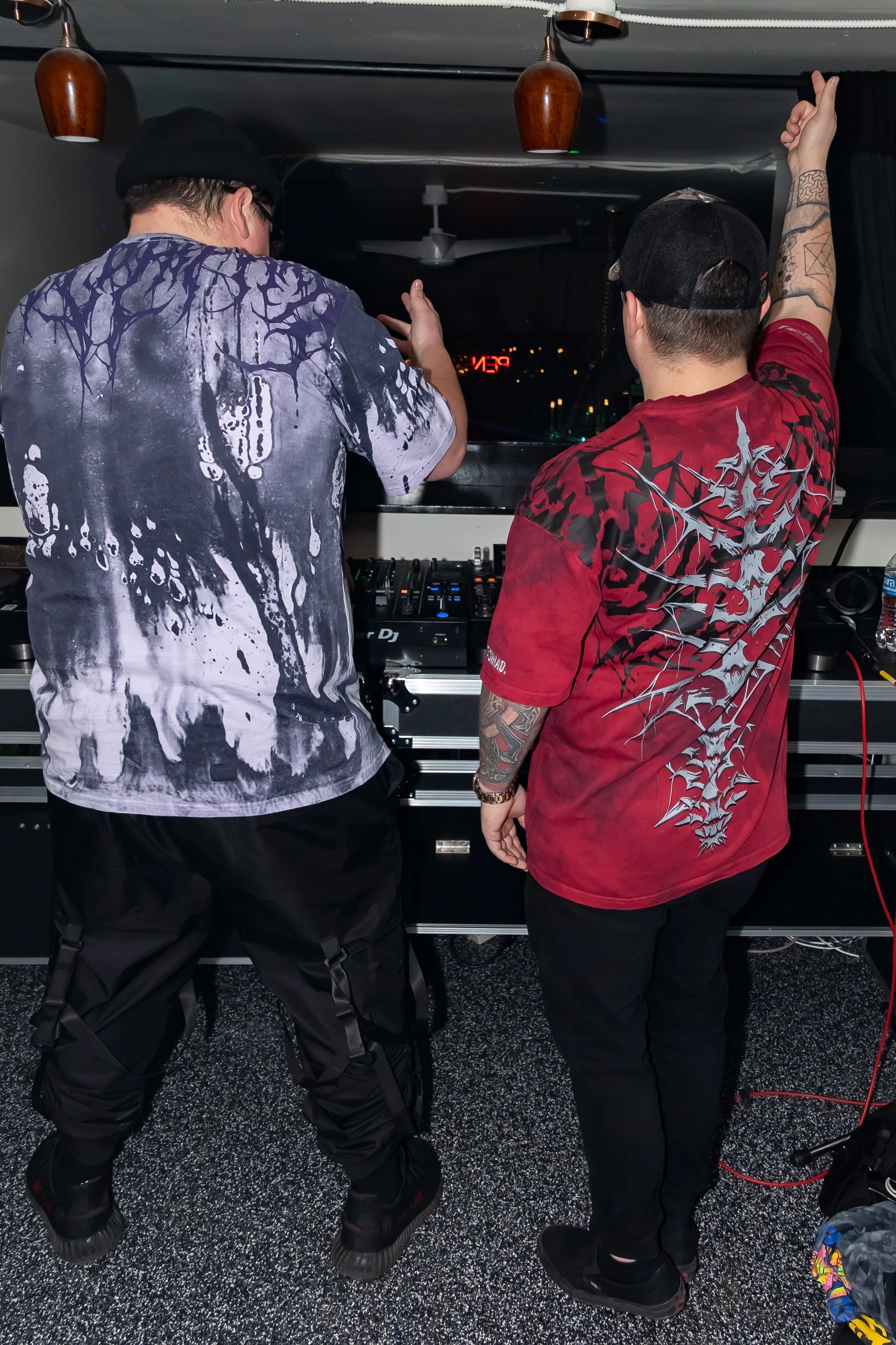Two DJs standing behind a DJ booth in a dimly lit room with dark curtains, ceiling fans, and hanging lights, facing away from the camera, wearing casual streetwear and caps, with a cityscape visible through a window in the background.