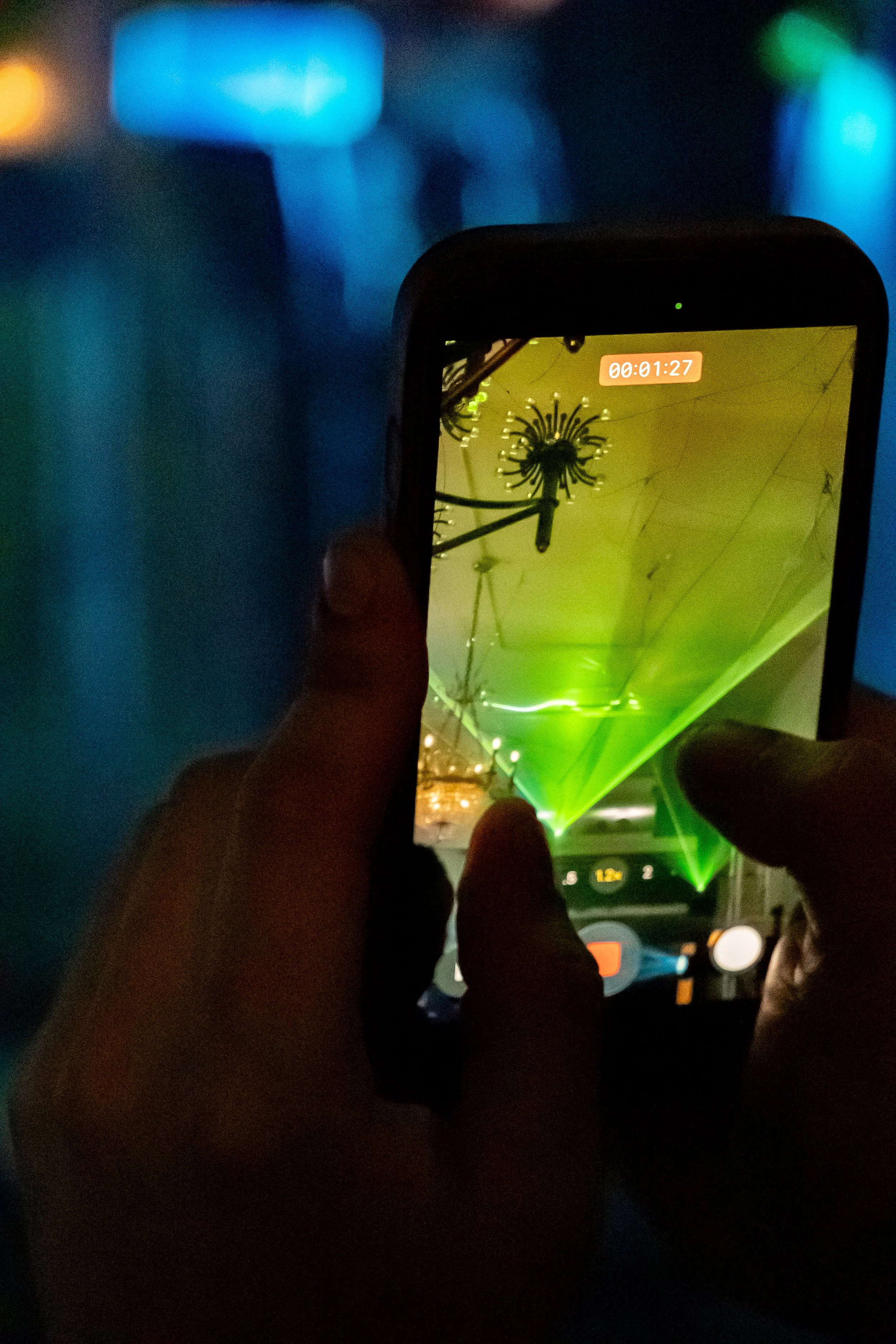 Person capturing a photo of a laser light show on a smartphone screen with green laser beams and spider webs on the ceiling.