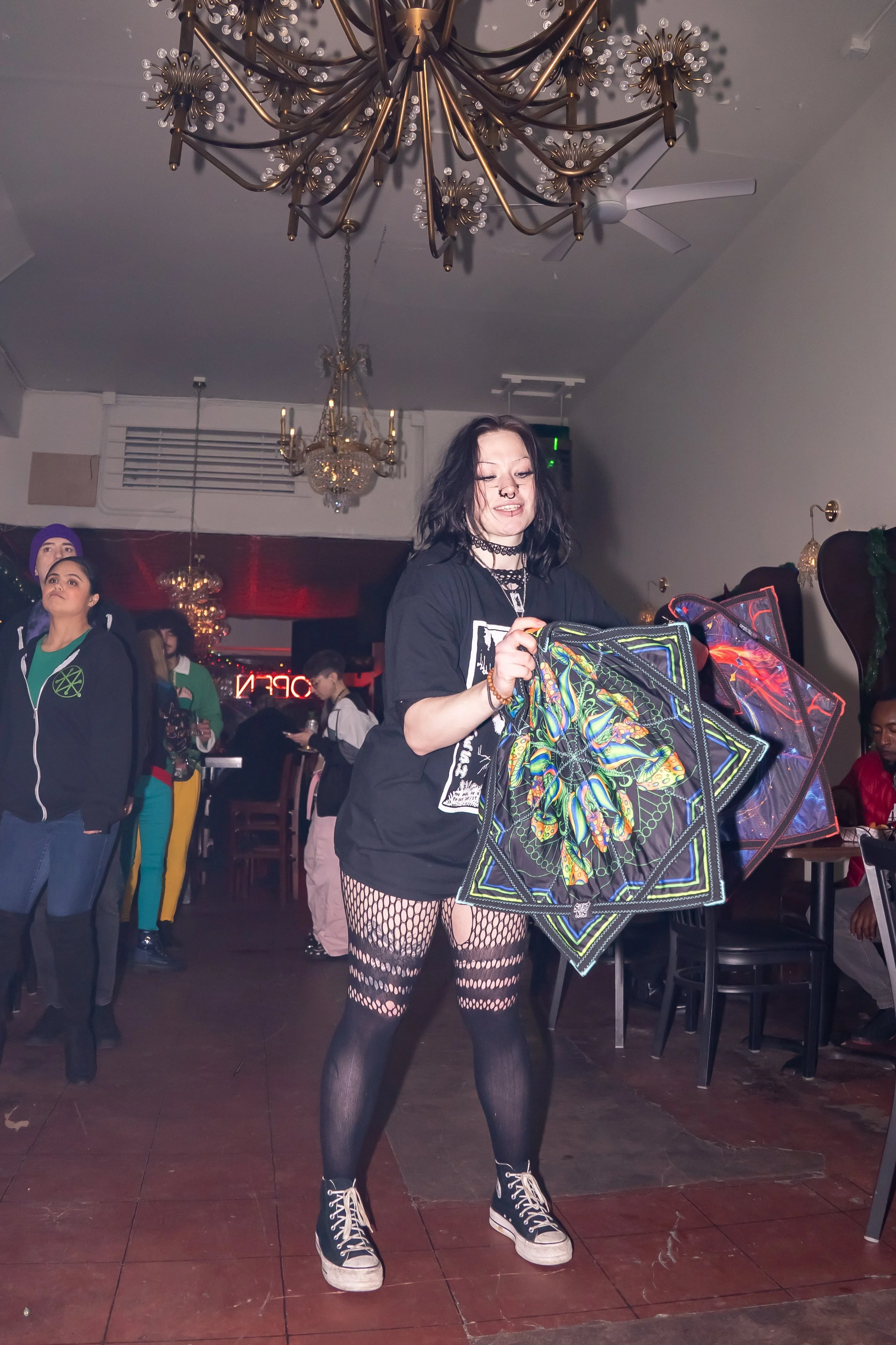 A young woman with black hair and fishnet stockings stands inside a restaurant or bar, holding colorful capes or cloths, with other people in the background. The restaurant has chandeliers on the ceiling and a neon sign.