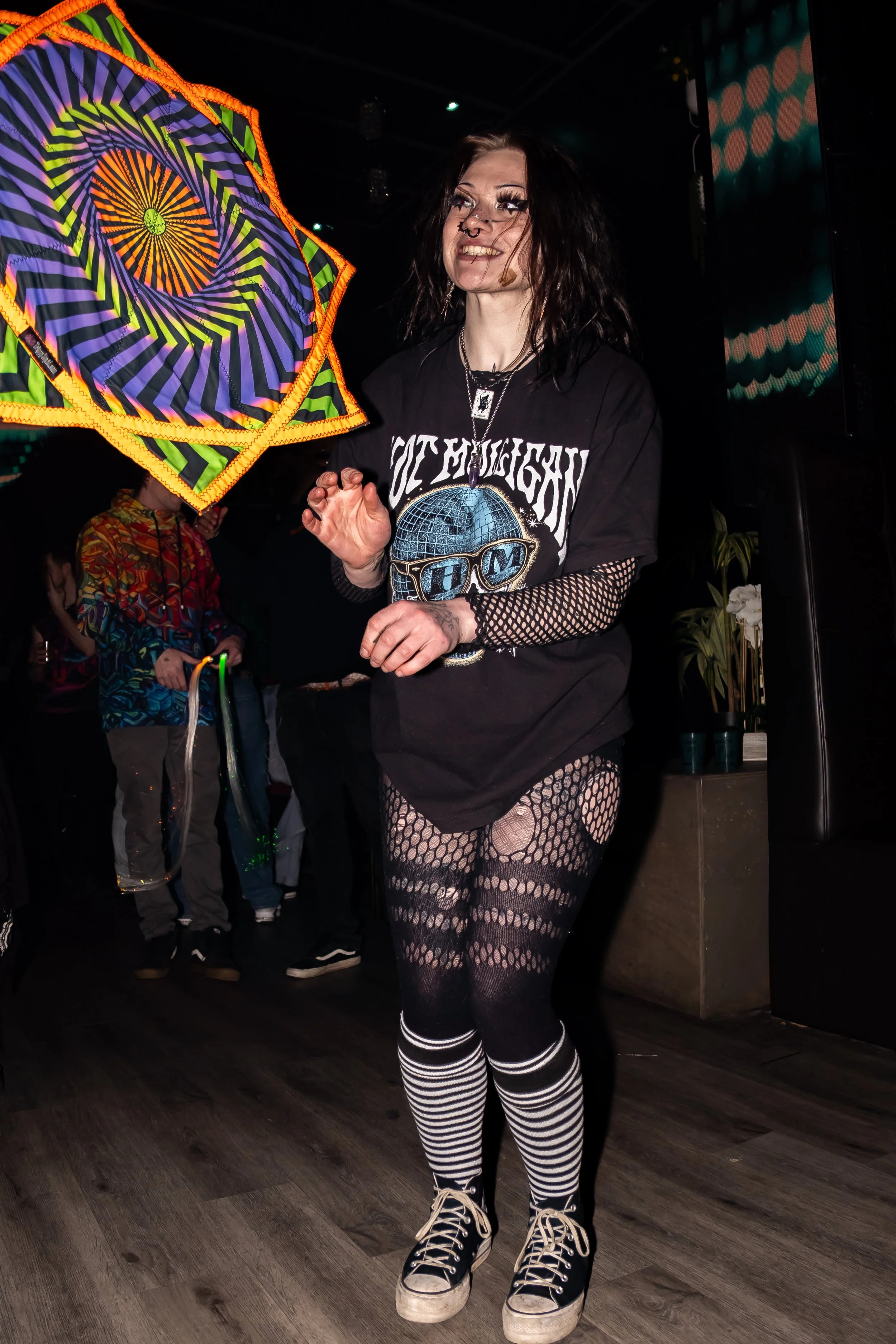A woman with black hair and fishnet clothing smiling at a party or concert, holding a colorful, illuminated umbrella.
