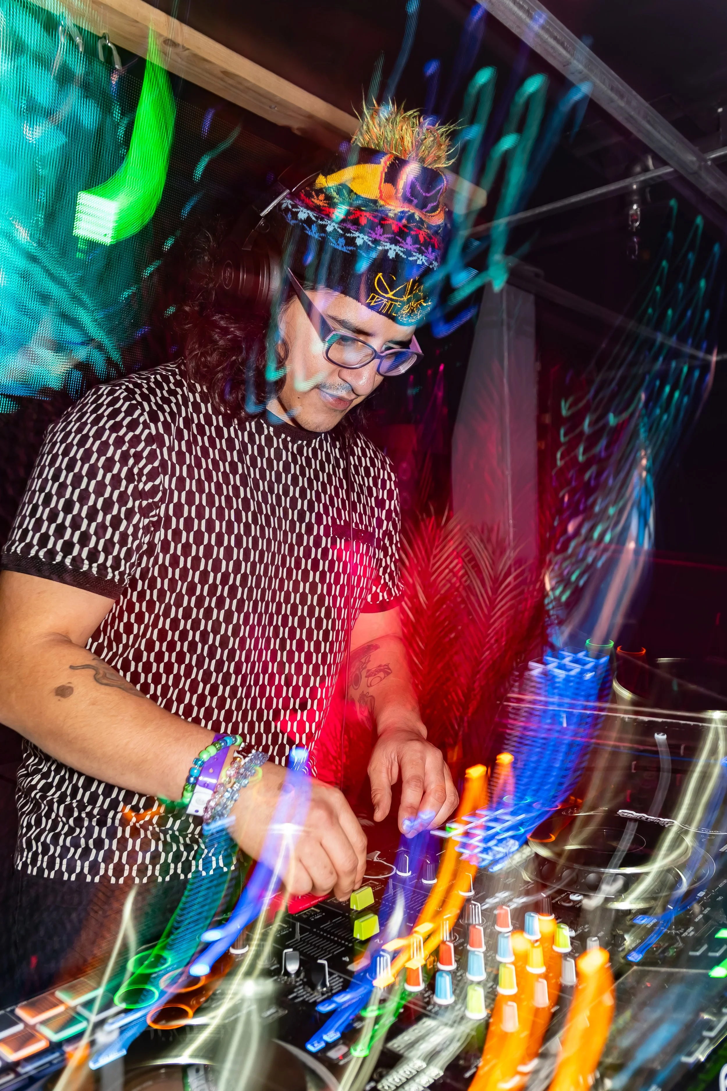 A DJ wearing glasses and a colorful beanie is mixing music on a turntable at a nightclub, with vibrant light effects creating streaks and patterns around him.