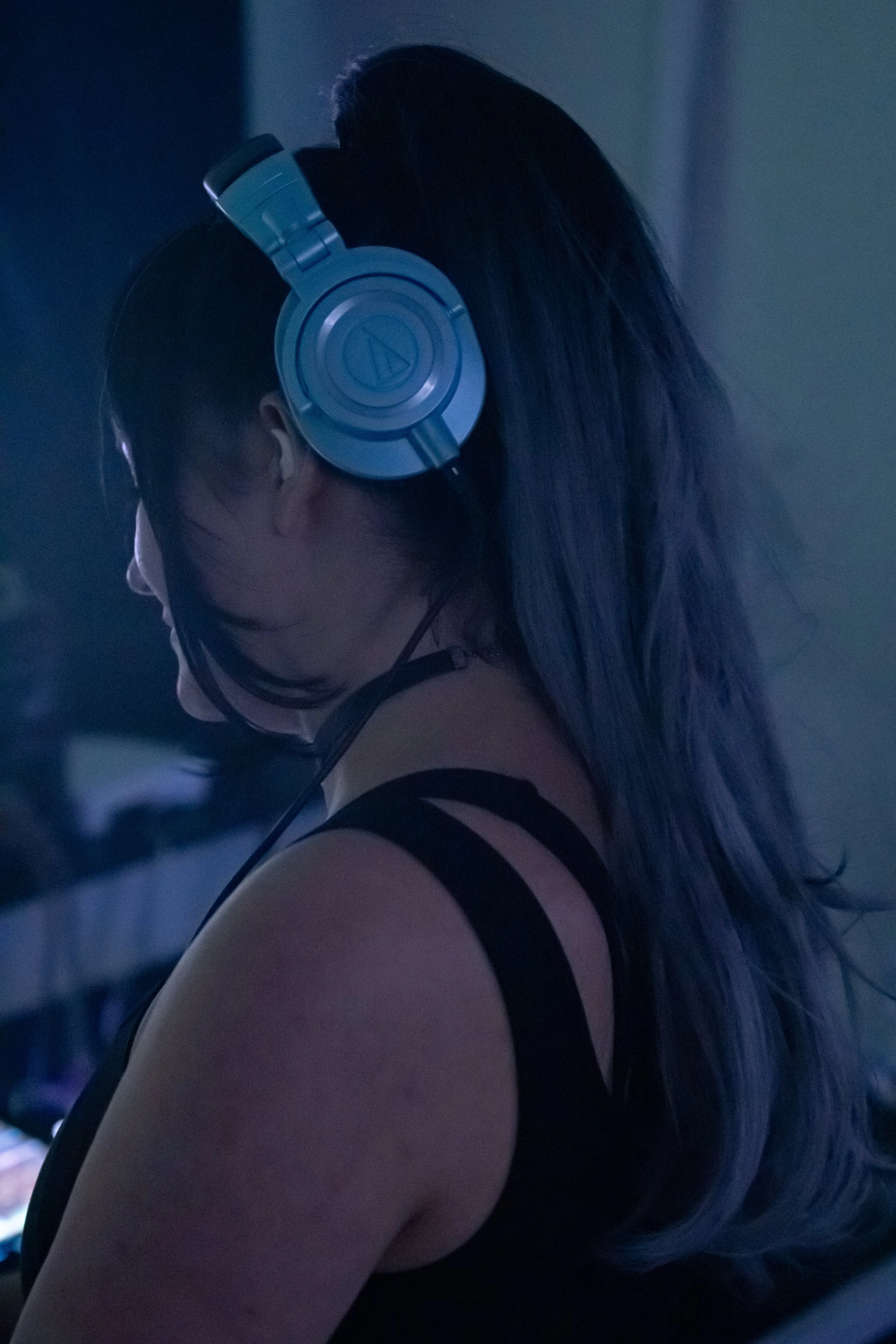 A woman with long hair wearing over-ear headphones, facing to the left, in a dimly lit indoor environment.