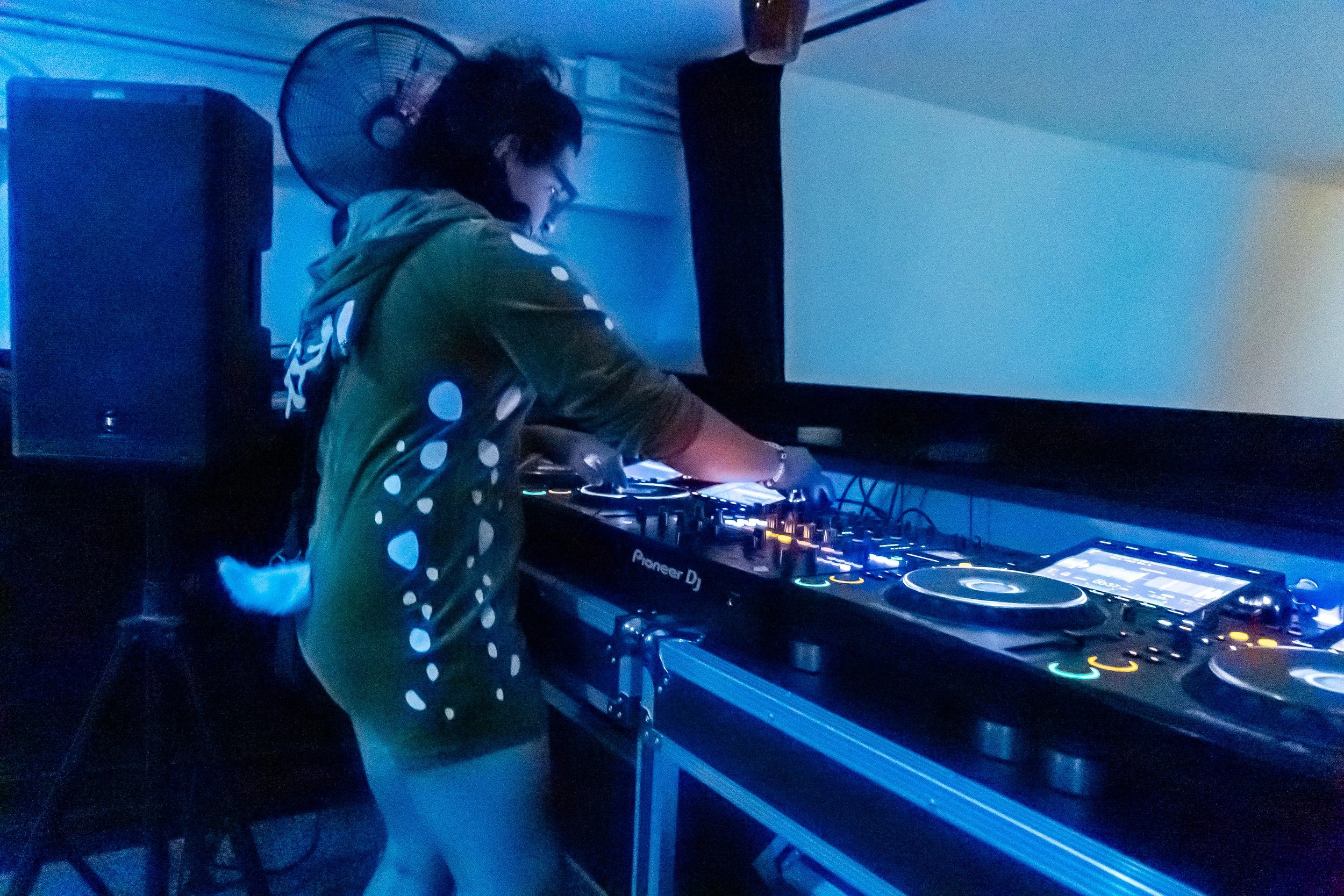 A person wearing a 3D cat tail and ears hoodie, DJing with Pioneer DJ equipment in a dimly lit room with blue lighting.