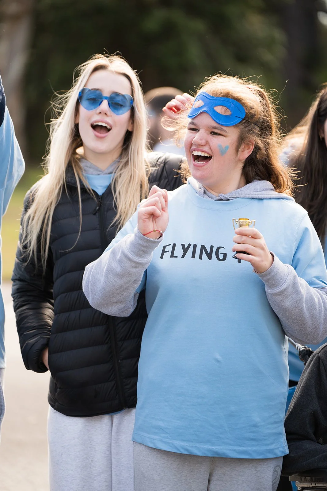 Two young women wearing blue masks and sunglasses and casual clothing, one holding a small trophy and celebrating outdoors.