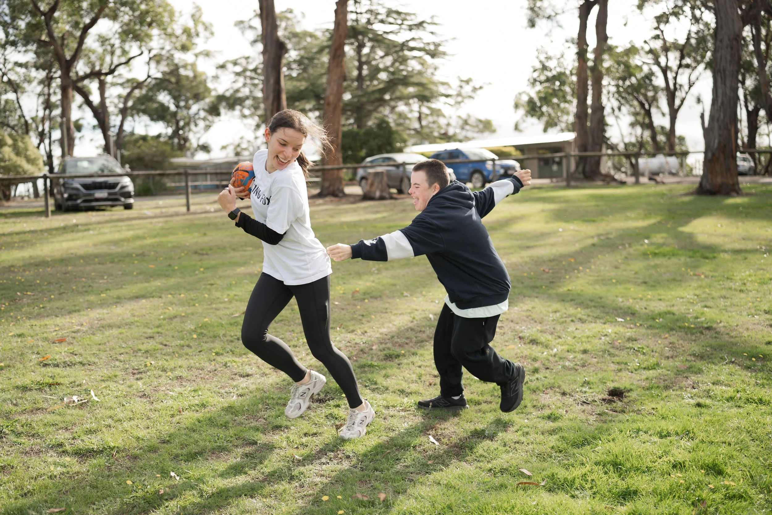 Two young people playing football on a grassy field with trees in the background, smiling and having fun.