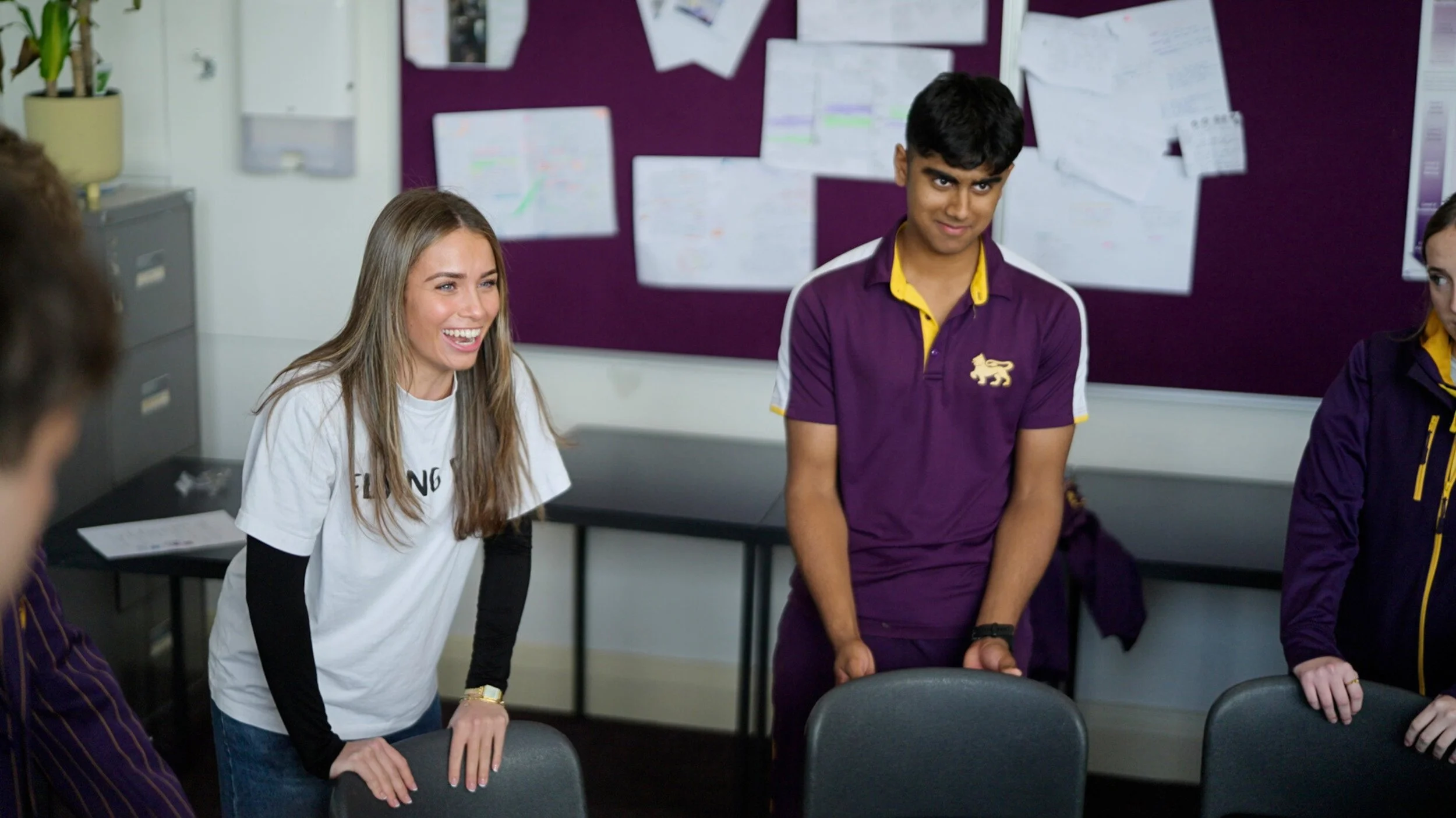 Group of students in a classroom, smiling and engaging in a discussion, with a purple bulletin board and papers on the wall in the background.