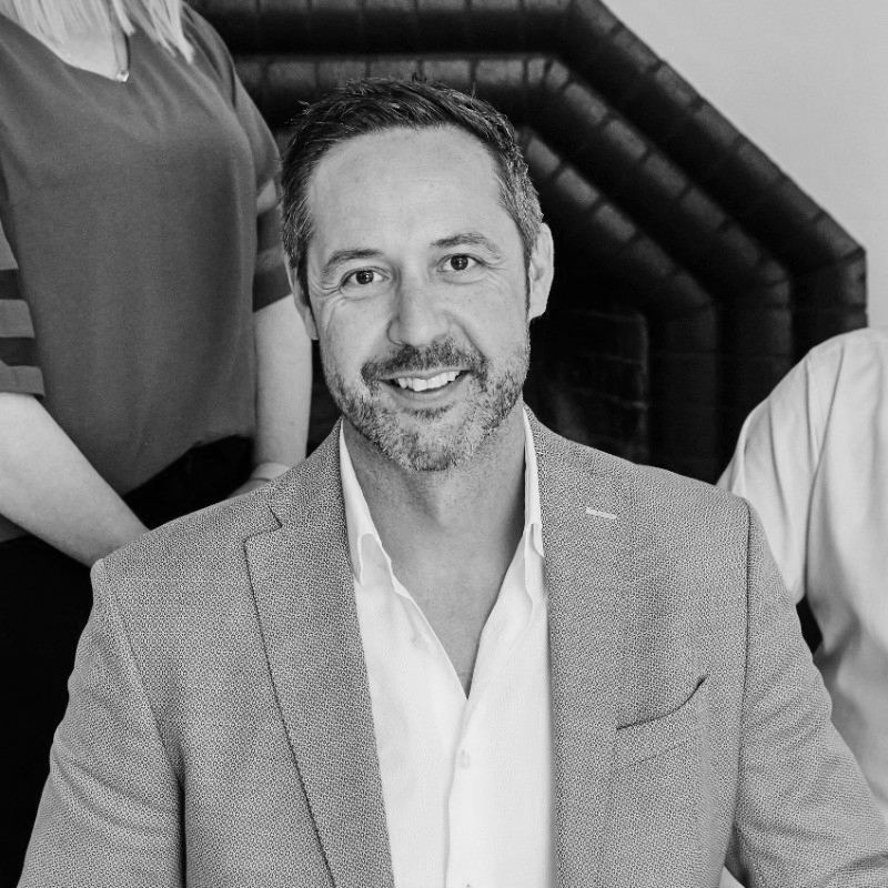 A smiling man with a beard and short hair, wearing a light-colored suit jacket and a white shirt, sitting indoors.
