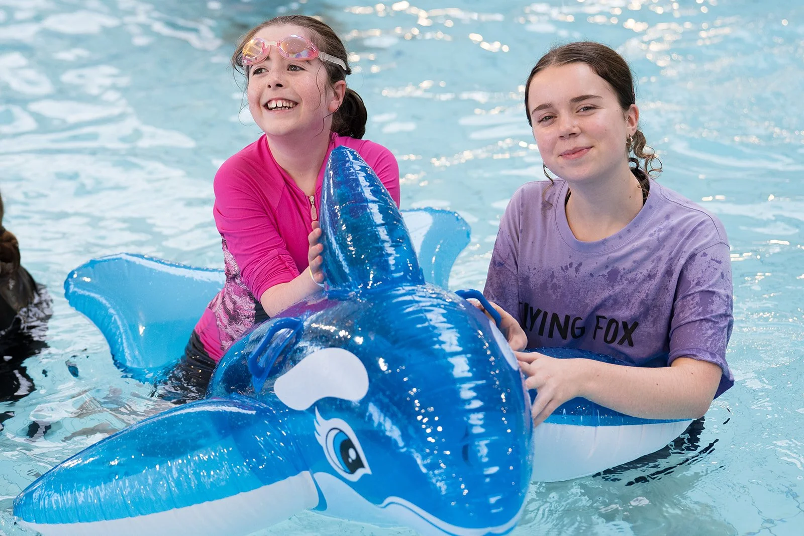 Two girls in a pool, one wearing pink and the other purple shirt, sitting on a blue inflatable shark float, smiling.
