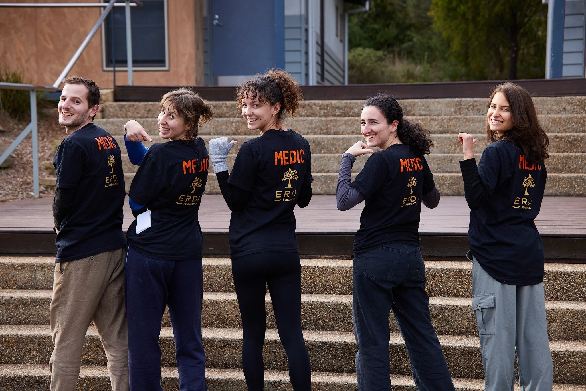 Group of five young emergency medical responders standing outdoors, facing backward, flexing their arms, wearing black T-shirts with 'MEDIC' printed in orange on the back, smiling, with a building and trees in the background.