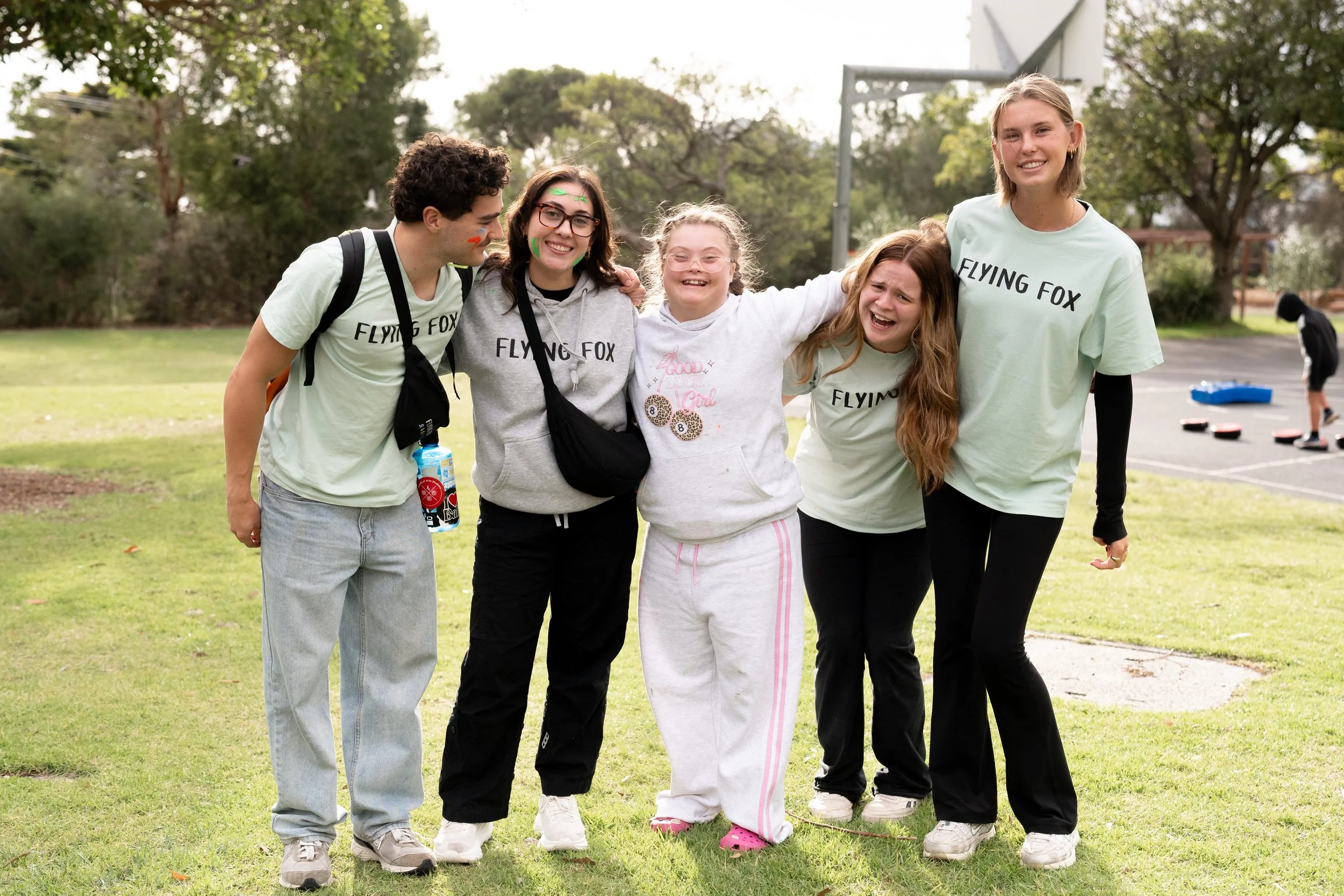 Group of five young people smiling and standing arm-in-arm outdoors in a park on a cloudy day, with trees and a basketball court in the background.