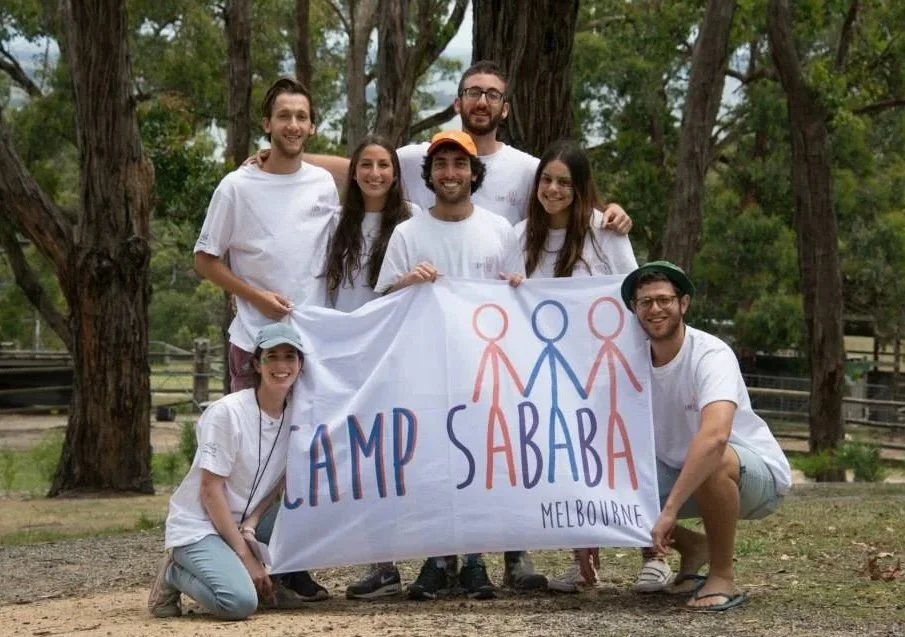 Group of seven young adults outdoors holding a Camp Sababa Melbourne banner, surrounded by trees, smiling and posing for a group photo.
