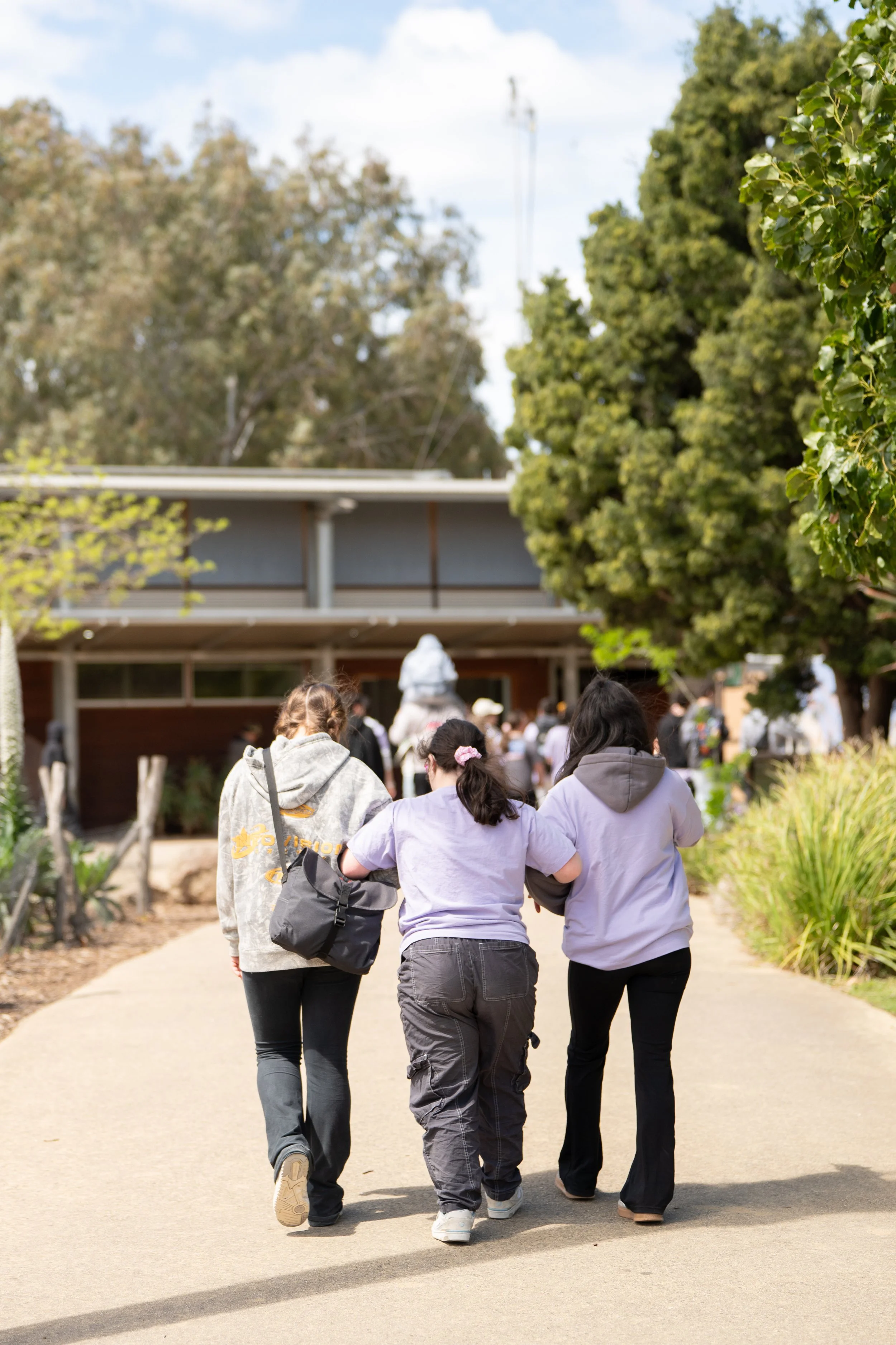 Three people walking on a sidewalk in a park-like area, heading towards a building, with trees and greenery around.
