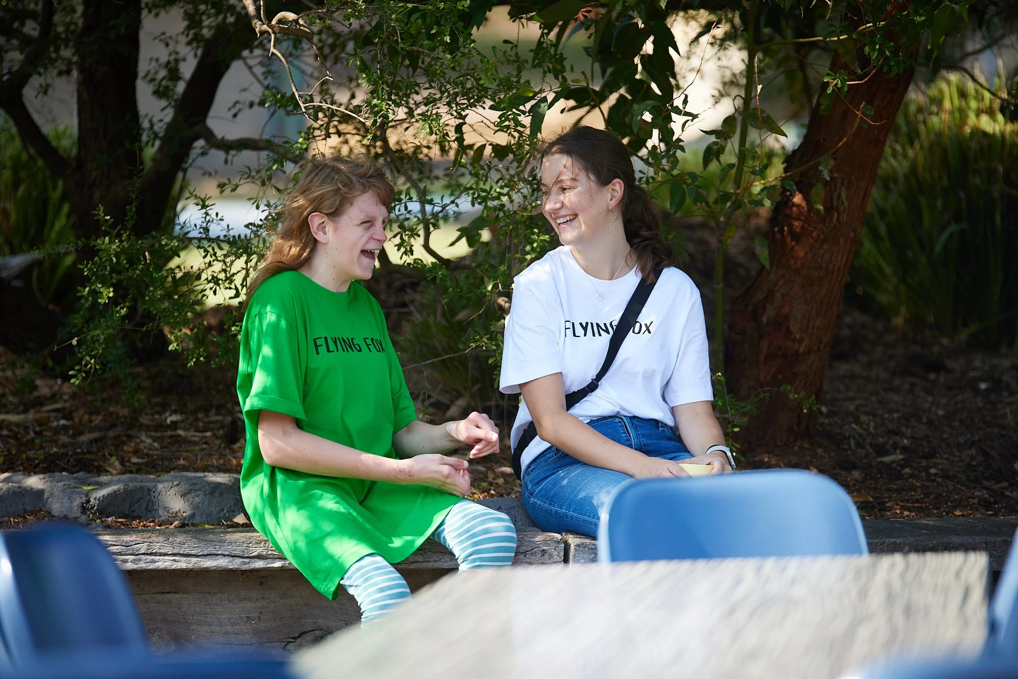 Two young women sitting outdoors in a park, laughing and talking. One wears a green t-shirt with 'Flying Fox' printed on it, and the other wears a white t-shirt with the same text.