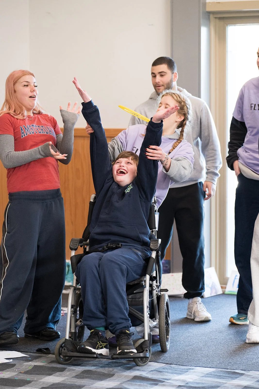 A group of young people, including a person in a wheelchair, are having fun indoors. One girl is throwing something in the air, and others are watching, smiling, and laughing.