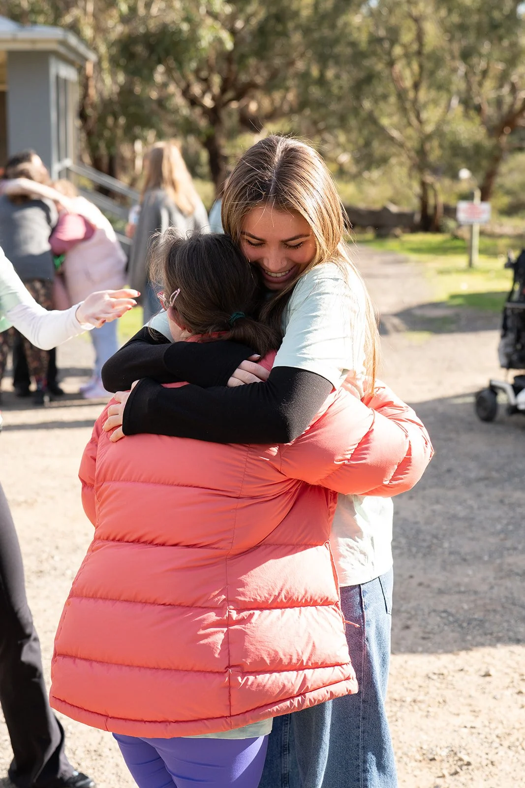 A young woman with long hair hugging a girl with glasses and dark hair outdoors on a sunny day. The young woman is smiling and appears happy. Several people are in the background, some hugging or talking, with trees and a building nearby.