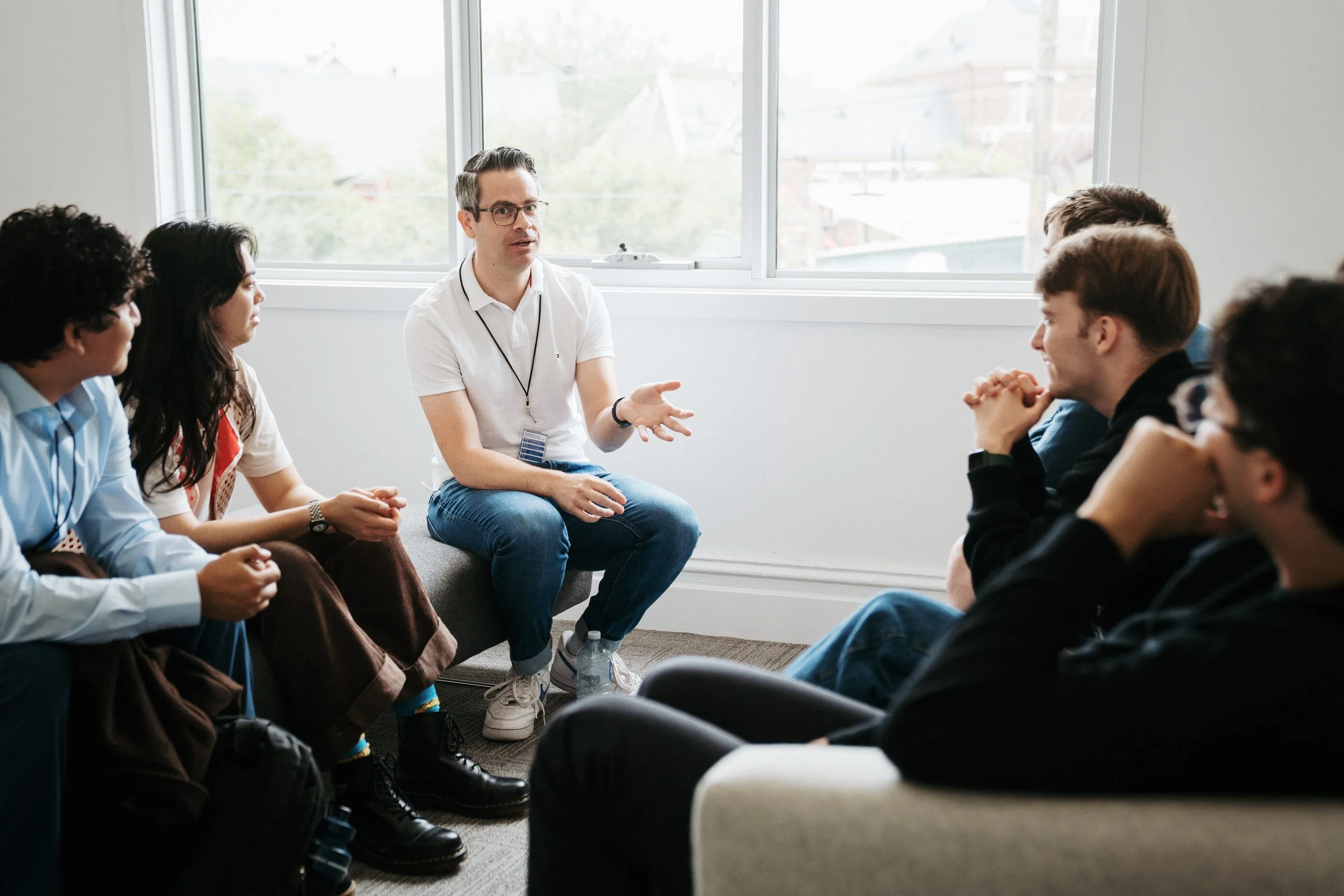 A man in a white shirt and glasses leading a discussion with a group of young people seated in a circle in a bright room with large window.