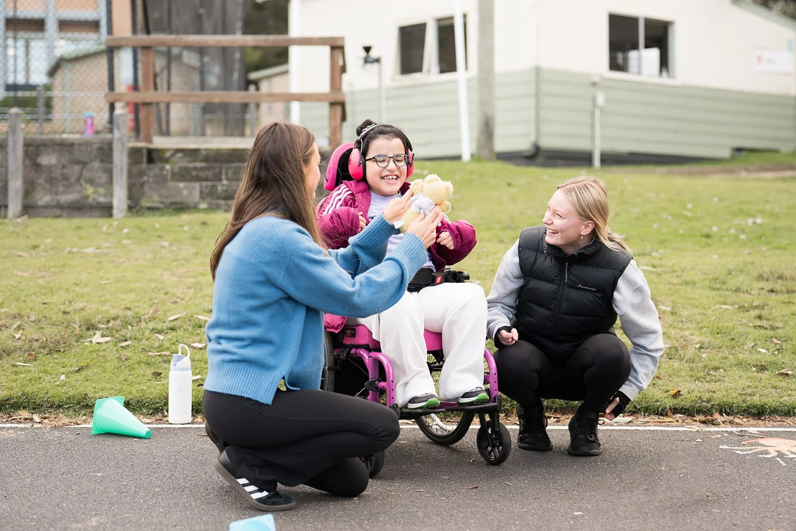 Three women are outdoors on a paved and grassy area, with one woman in a wheelchair holding a teddy bear, smiling, as the other two women crouch nearby, smiling and engaging with her. One is wearing a blue sweater and black pants, the other is in a black vest and gray hoodie. A water bottle and a small green cone are on the ground nearby.