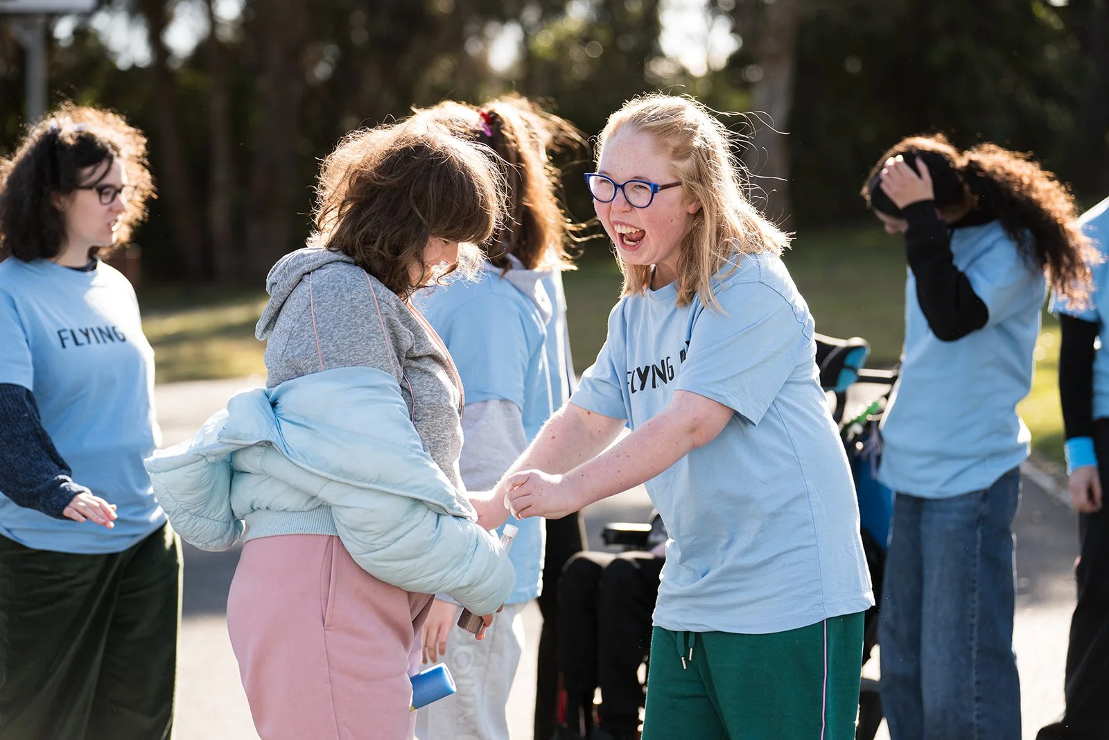 A group of young women outdoors, with one girl laughing and holding hands with another, while others are standing nearby on a sunny day.