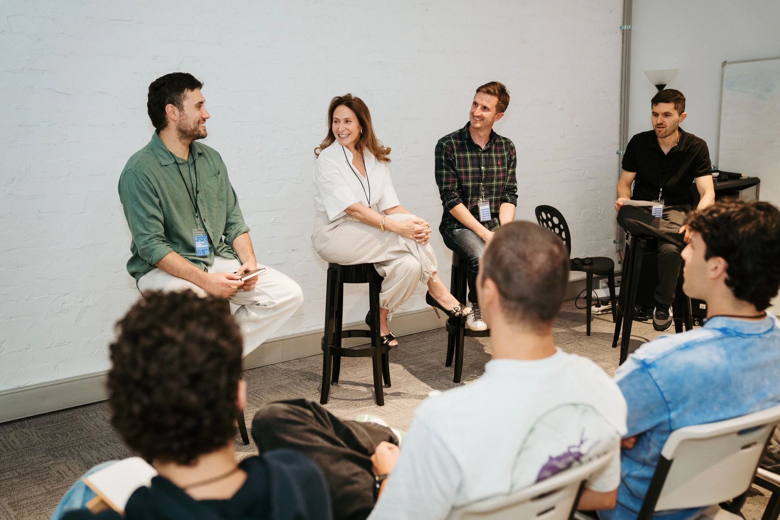 Panel discussion in a room with five people on stage, two women and three men, engaging with audience members who are seated. The panelists are sitting on chairs in front of a white brick wall, and some are holding microphones. Audience members are listening attentively.