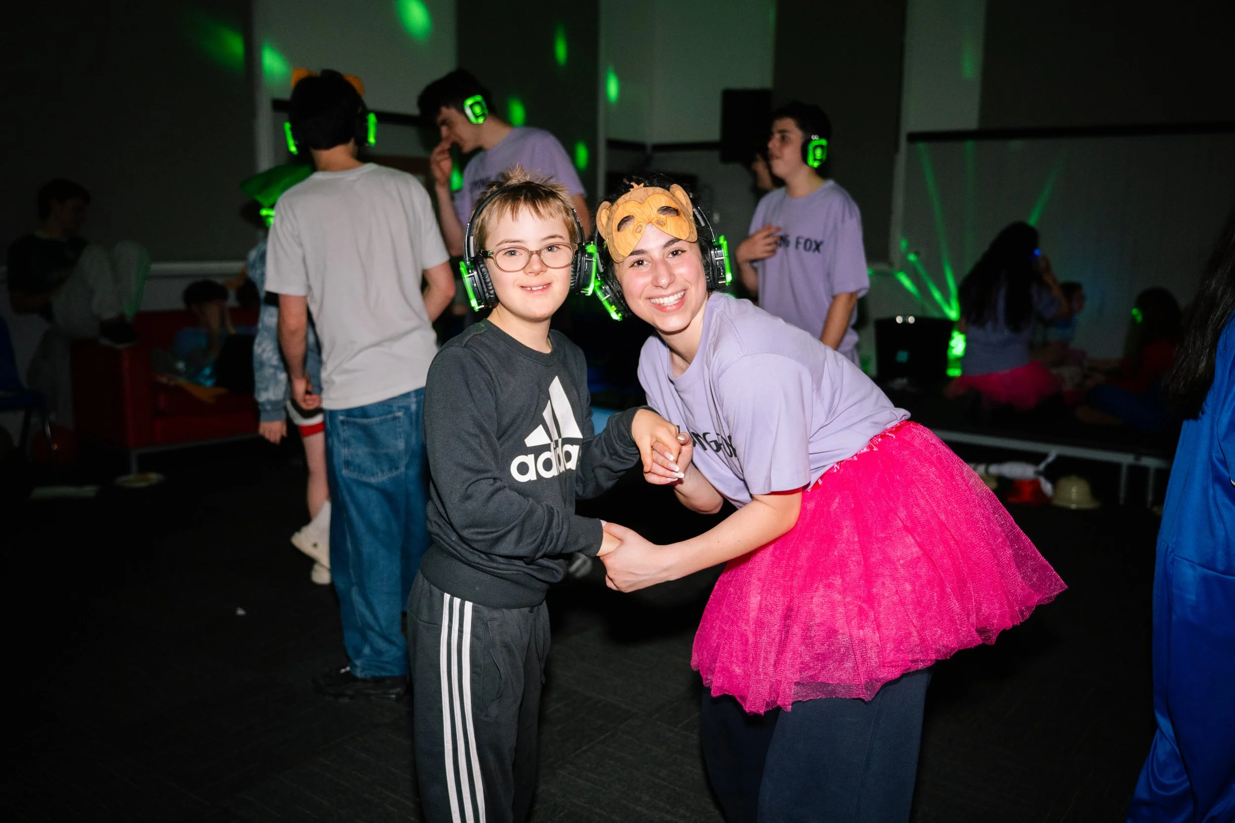 A woman and a boy, both wearing headphones, holding hands and smiling at the camera on a dance floor. The woman is wearing a purple shirt, pink tutu, and a mask on her head. Several other kids are in the background with headphones and some with ears or hats.